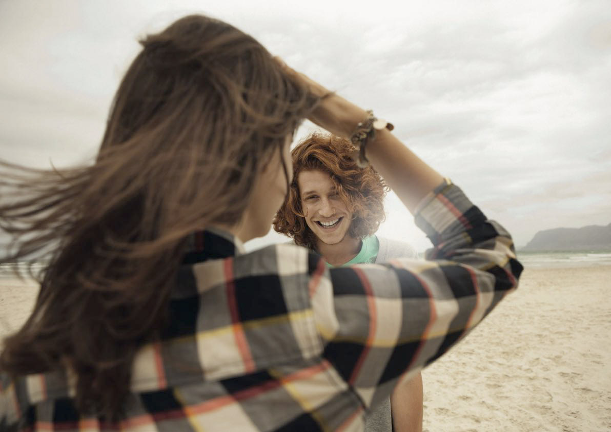 Ein fröhlicher junger Mann mit lockigem Haar lächelt eine Frau an einem bewölkten Strandtag breit an, während sie spielerisch seine Haare zurechtrückt. Beide genießen einen unbeschwerten Moment. © Fotografie Tomas Rodriguez