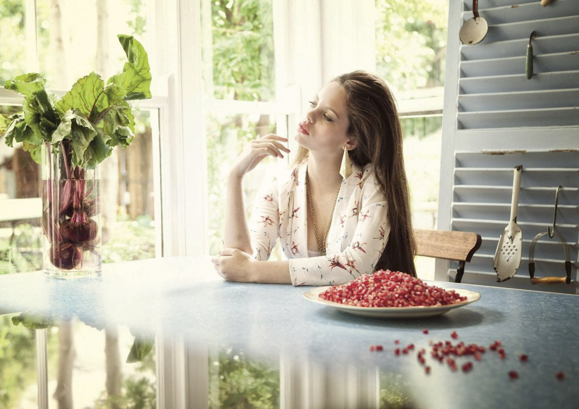 Eine Frau sitzt gedankenverloren an einem Küchentisch und blickt aus dem Fenster. Auf dem Tisch liegen verstreut rote Johannisbeeren und eine Vase mit Laub verleiht der Szene einen Hauch von Grün. © Fotografie Tomas Rodriguez