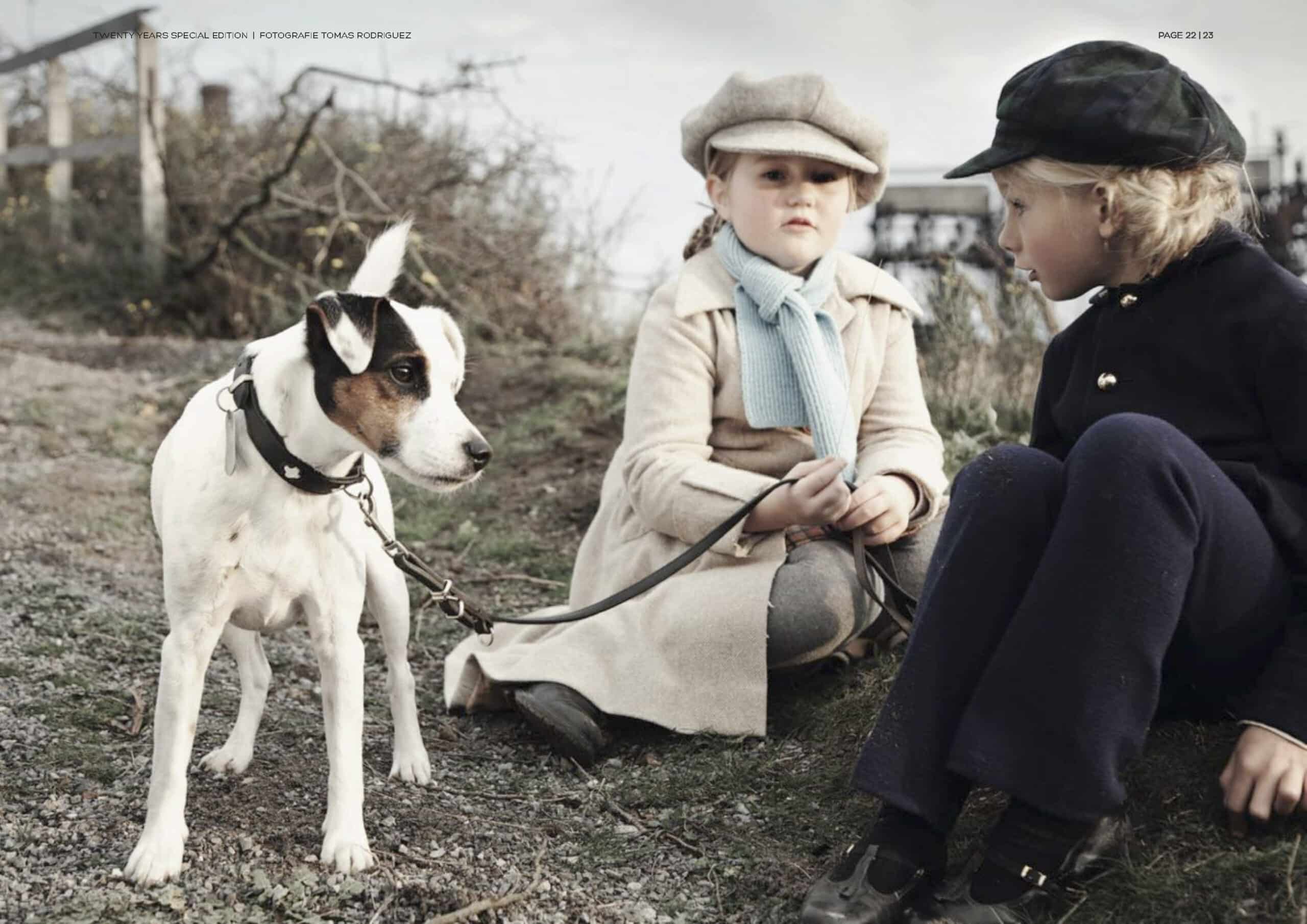 Zwei kleine Kinder in Vintage-Kleidung sitzen auf einem grasbewachsenen Hügel und neben ihnen ist ein weiß-brauner Hund an der Leine. Dies stellt eine heitere, altmodische Szene dar. Ein Kind starrt das andere intensiv an. © Fotografie Tomas Rodriguez