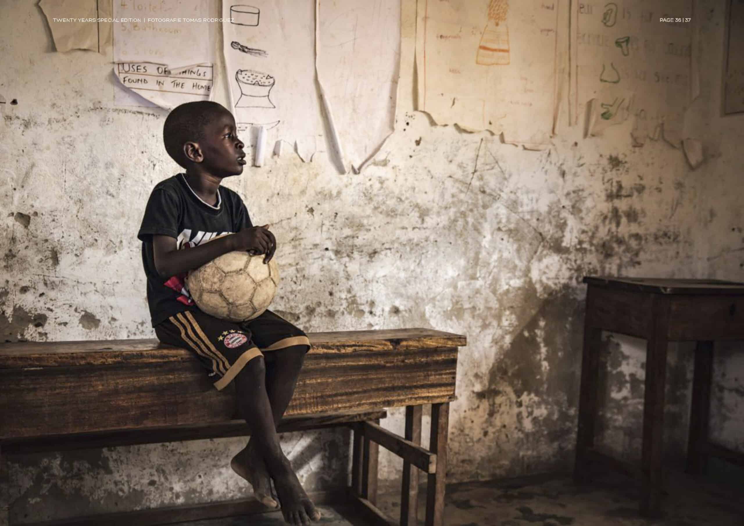 Ein kleiner Junge sitzt mit einem Fußball in der Hand auf einer Holzbank in einem Klassenzimmer mit Zeichnungen und Unterrichtsmaterialien an den Wänden. © Fotografie Tomas Rodriguez