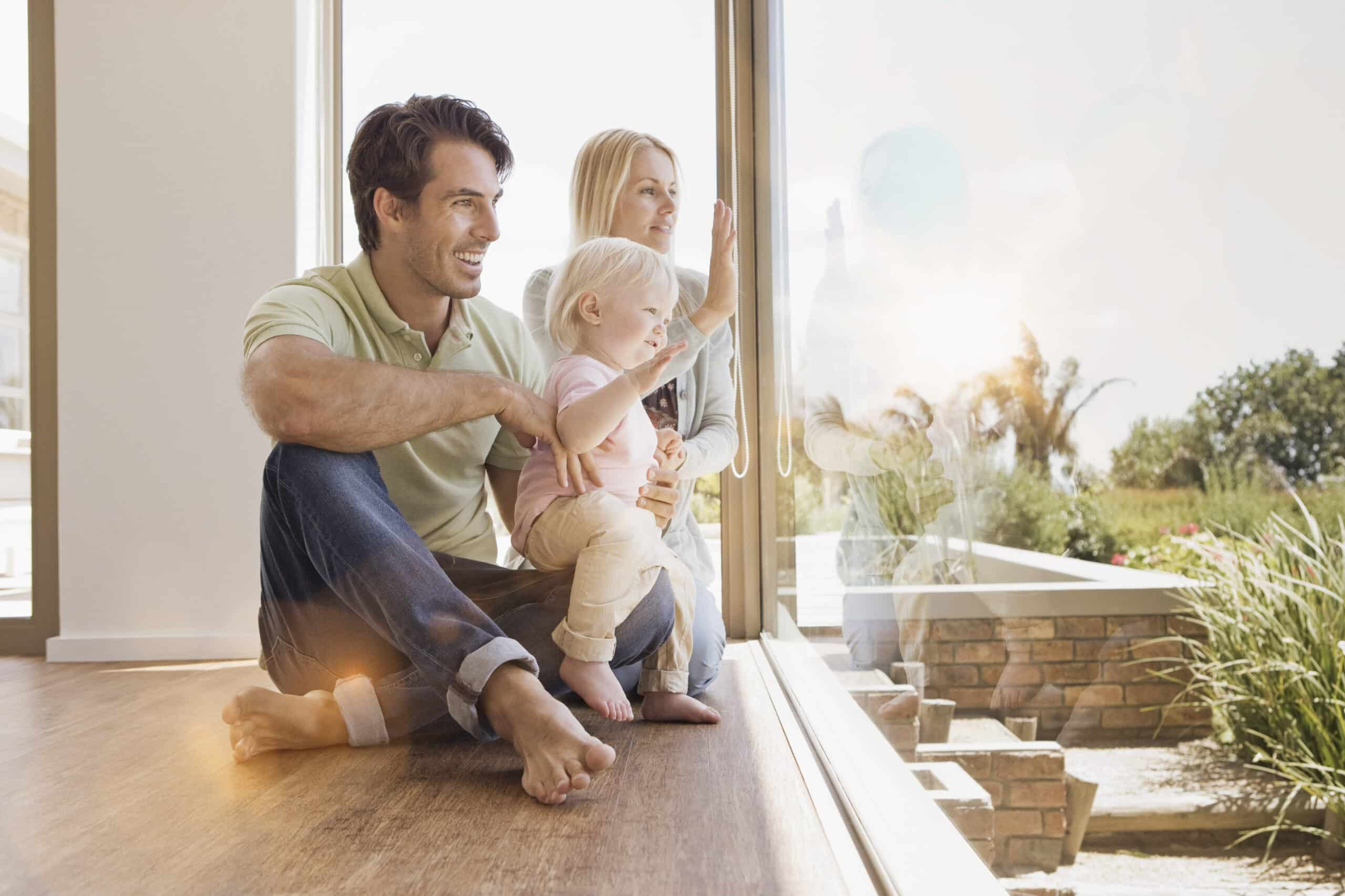 Eine dreiköpfige Familie, bestehend aus Vater, Mutter und kleinem Kind, sitzt an einem großen Fenster mit malerischer Aussicht. Vater und Kind sitzen auf dem Boden, während die Mutter hinter ihnen steht. Sie alle wirken entspannt und glücklich, während sie nach draußen blicken. © Fotografie Tomas Rodriguez