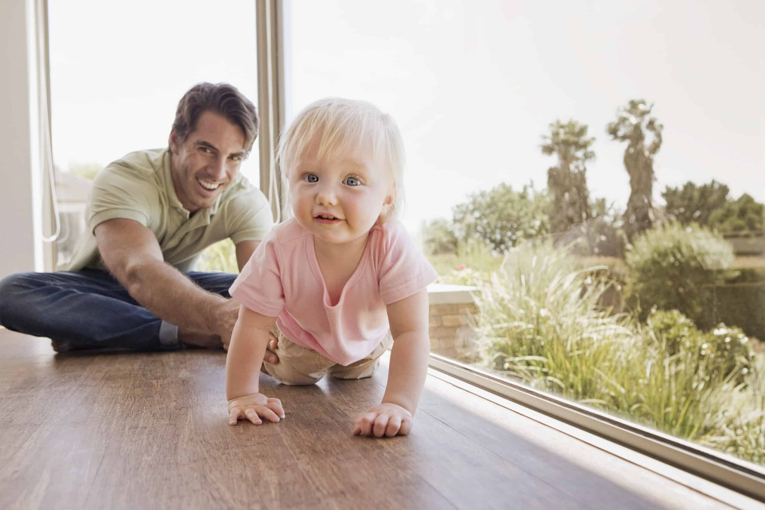 Ein lächelnder Mann, der auf dem Boden sitzt, streckt die Hand nach einem Baby aus, das auf einem Holzboden neben großen Fenstern krabbelt. Das Baby, das ein rosa Hemd trägt, schaut nach vorne. Der Hintergrund zeigt eine sonnige Szene im Freien mit viel Grün und Palmen. © Fotografie Tomas Rodriguez