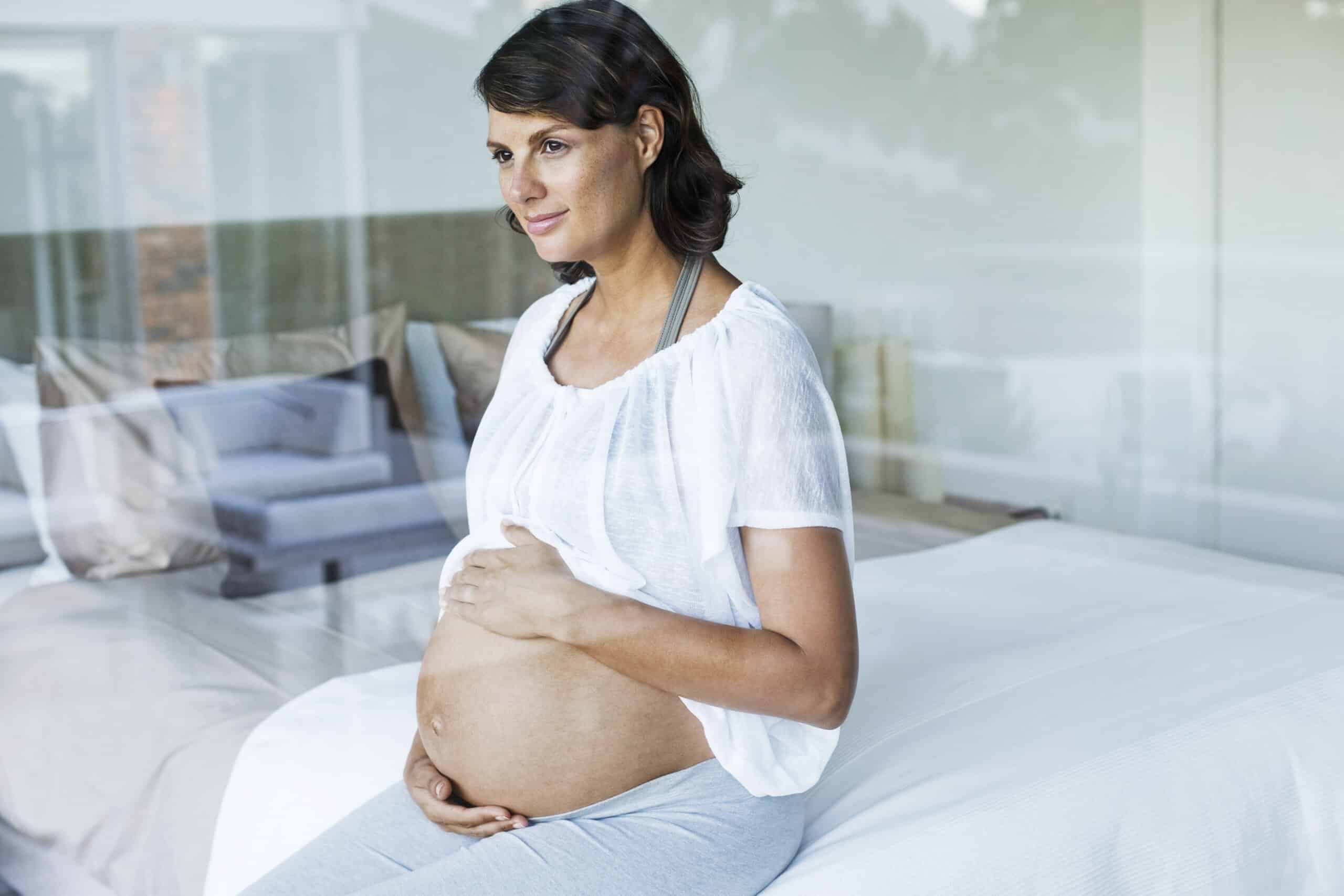 Eine schwangere Frau sitzt auf einem Bett und hält mit beiden Händen sanft ihren Babybauch. Sie hat kurze, dunkle Haare und trägt ein weißes, kurzärmliges Oberteil und eine graue Hose. Im Hintergrund ist ein heller, moderner Raum mit einem Sofa und großen Fenstern zu sehen. © Fotografie Tomas Rodriguez