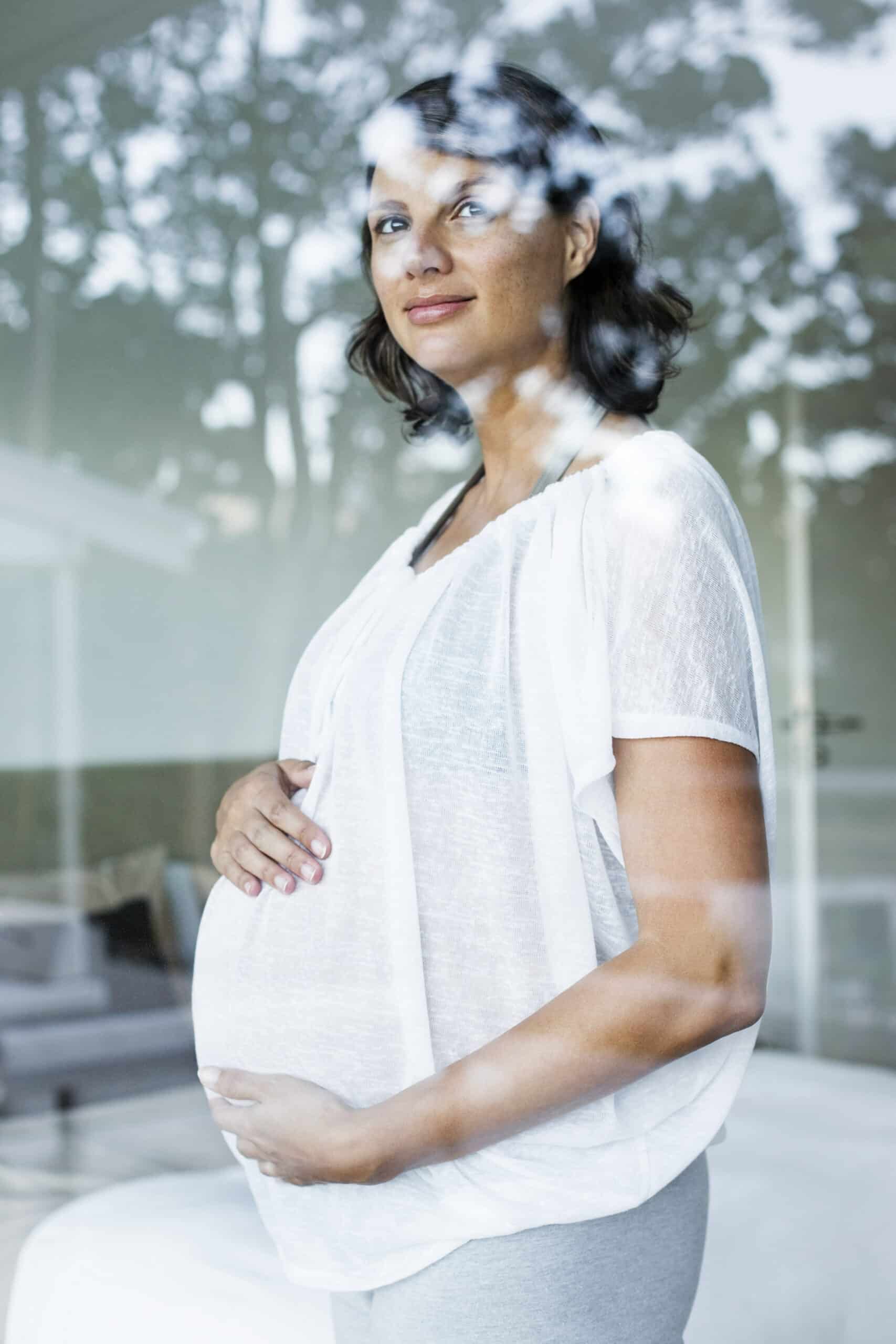 Eine schwangere Frau mit schulterlangem dunklem Haar blickt nachdenklich aus einem Fenster. Sie trägt eine weiße Bluse und hellgraue Hosen und hält ihren Bauch mit beiden Händen. Im Hintergrund ist eine verschwommene Innenkulisse zu sehen, in deren Glas sich Bäume spiegeln. © Fotografie Tomas Rodriguez