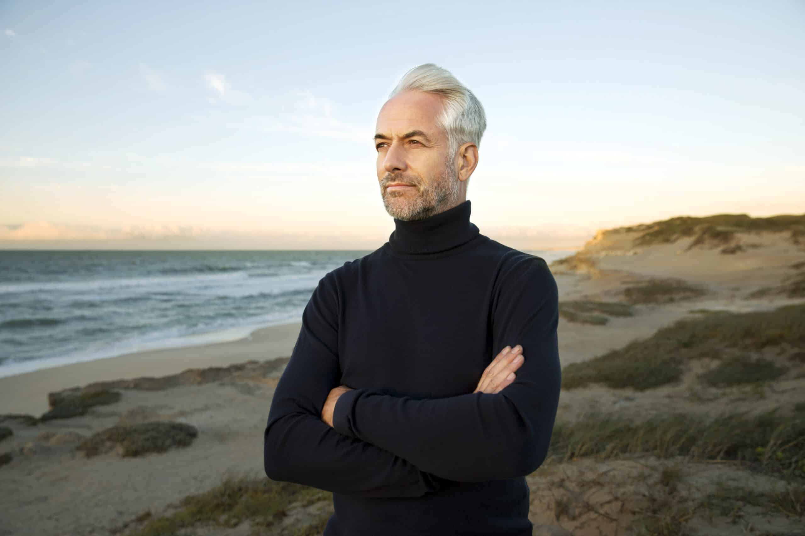 Ein Mann mit grauem Haar steht mit verschränkten Armen an einem Strand und trägt einen schwarzen Rollkragenpullover. Hinter ihm sind der Sandstrand und das blaue Meer unter einem klaren Himmel zu sehen. Sein Gesichtsausdruck wirkt nachdenklich, während er in die Ferne blickt. © Fotografie Tomas Rodriguez