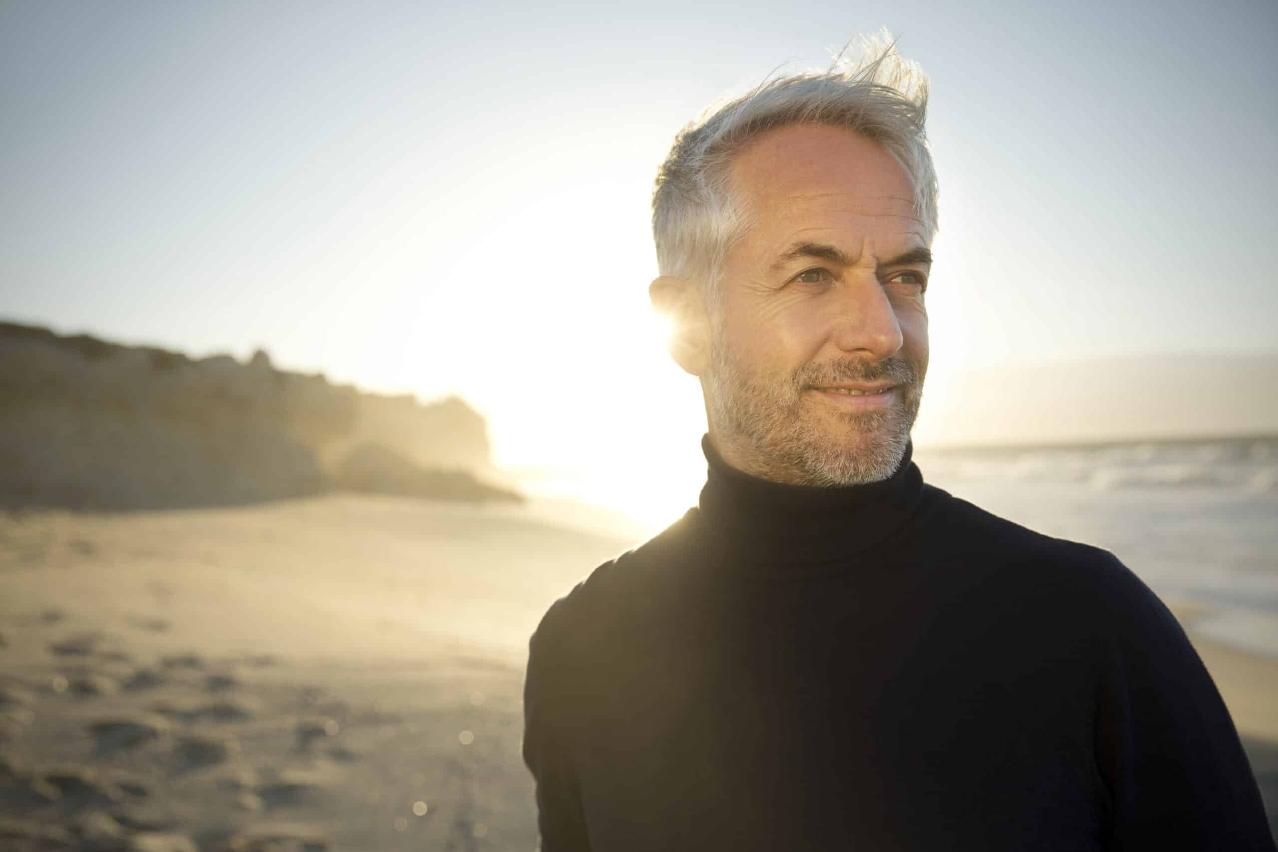 Ein Mann mit grauem Haar und Bart steht bei Sonnenuntergang an einem Sandstrand. Er trägt einen schwarzen Rollkragenpullover und blickt in die Ferne, während die Sonne hell hinter ihm scheint. Im Hintergrund sind Wellen und Klippen zu sehen. © Fotografie Tomas Rodriguez