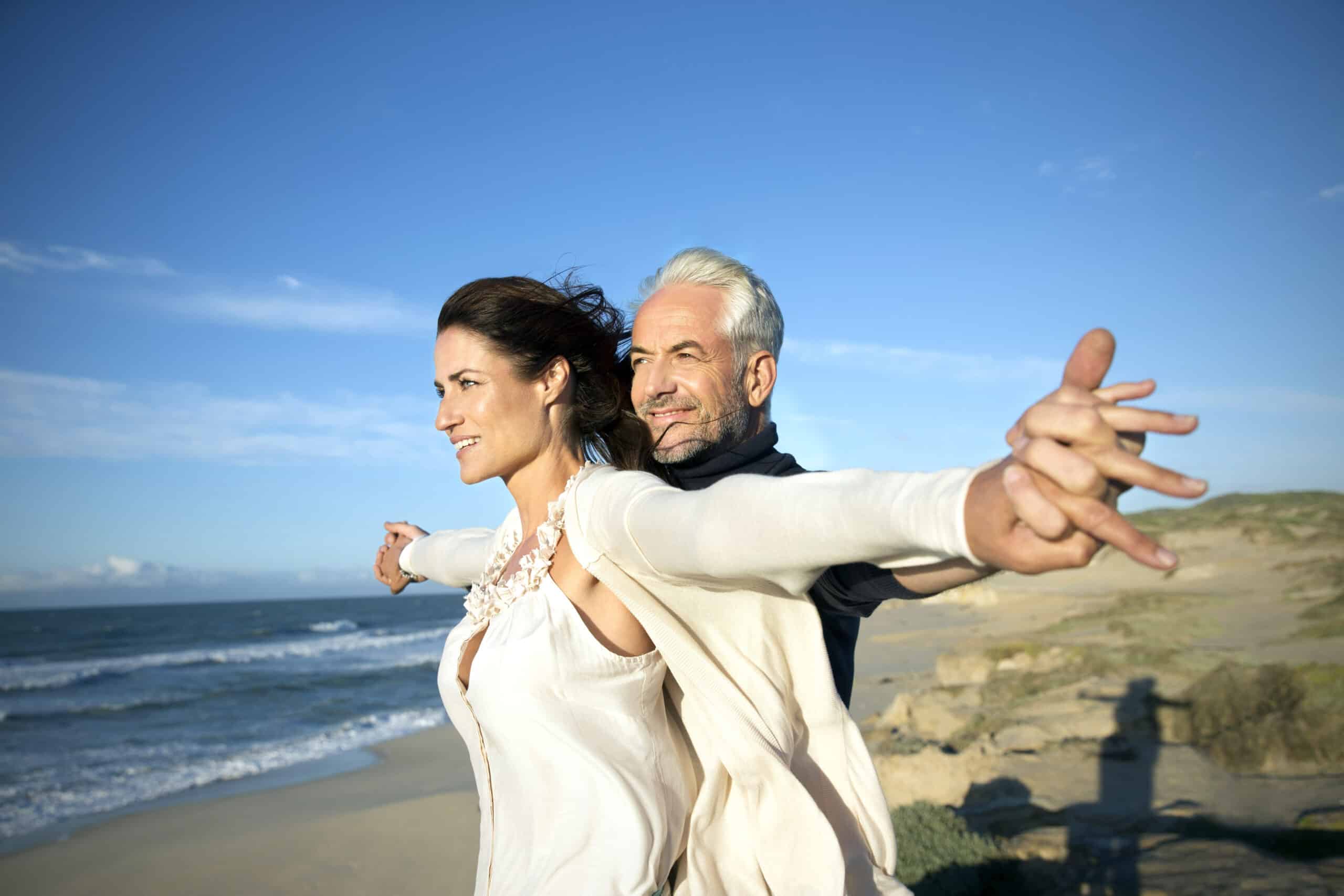 Ein lächelndes Paar steht mit ausgestreckten Armen am Strand und blickt auf das Meer. Die Frau steht vor ihr, der Mann steht hinter ihr und hält sie an der Taille. Sie scheinen die Brise und das Sonnenlicht zu genießen. Der Himmel ist klar mit ein paar Wolken. © Fotografie Tomas Rodriguez