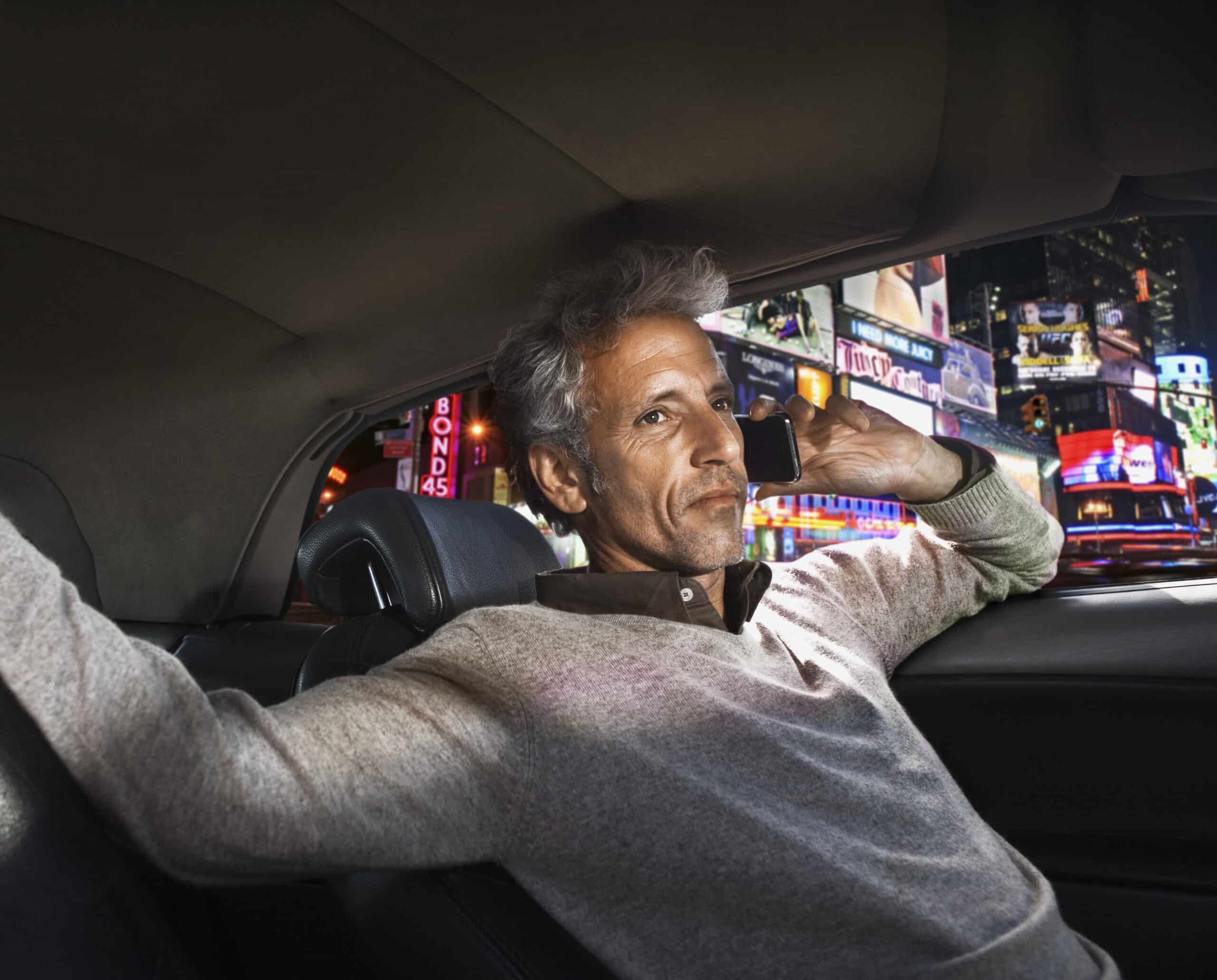 Ein Mann mit grauem Haar sitzt nachts auf dem Rücksitz eines Autos und hält ein Smartphone an sein Ohr. Er wirkt entspannt, ein Arm ruht auf der Rückenlehne des Sitzes. Durch das Autofenster sind leuchtende Lichter der Stadt und Werbetafeln zu sehen. © Fotografie Tomas Rodriguez