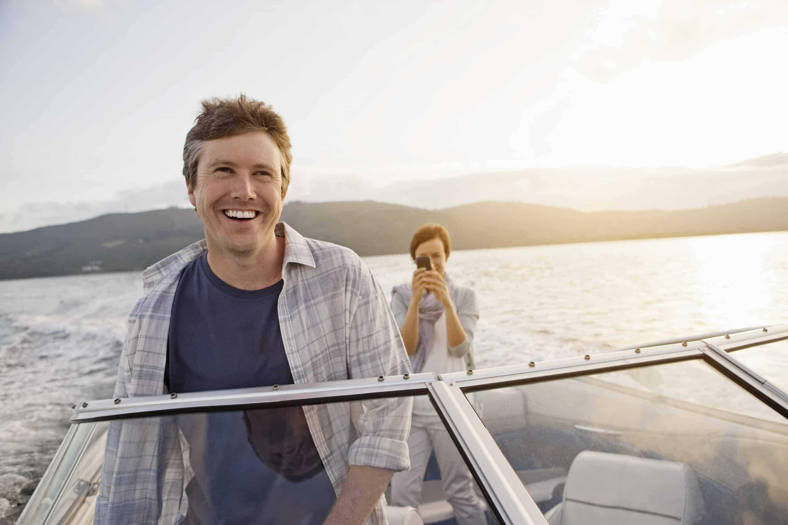 Ein Mann mit kurzen Haaren und legerem Hemd lächelt und fährt ein Schnellboot. Hinter ihm steht eine andere Person, hält ein Smartphone und macht ein Foto. Sie sind auf einem See oder dem Meer mit Bergen im Hintergrund und die Sonne geht unter. © Fotografie Tomas Rodriguez