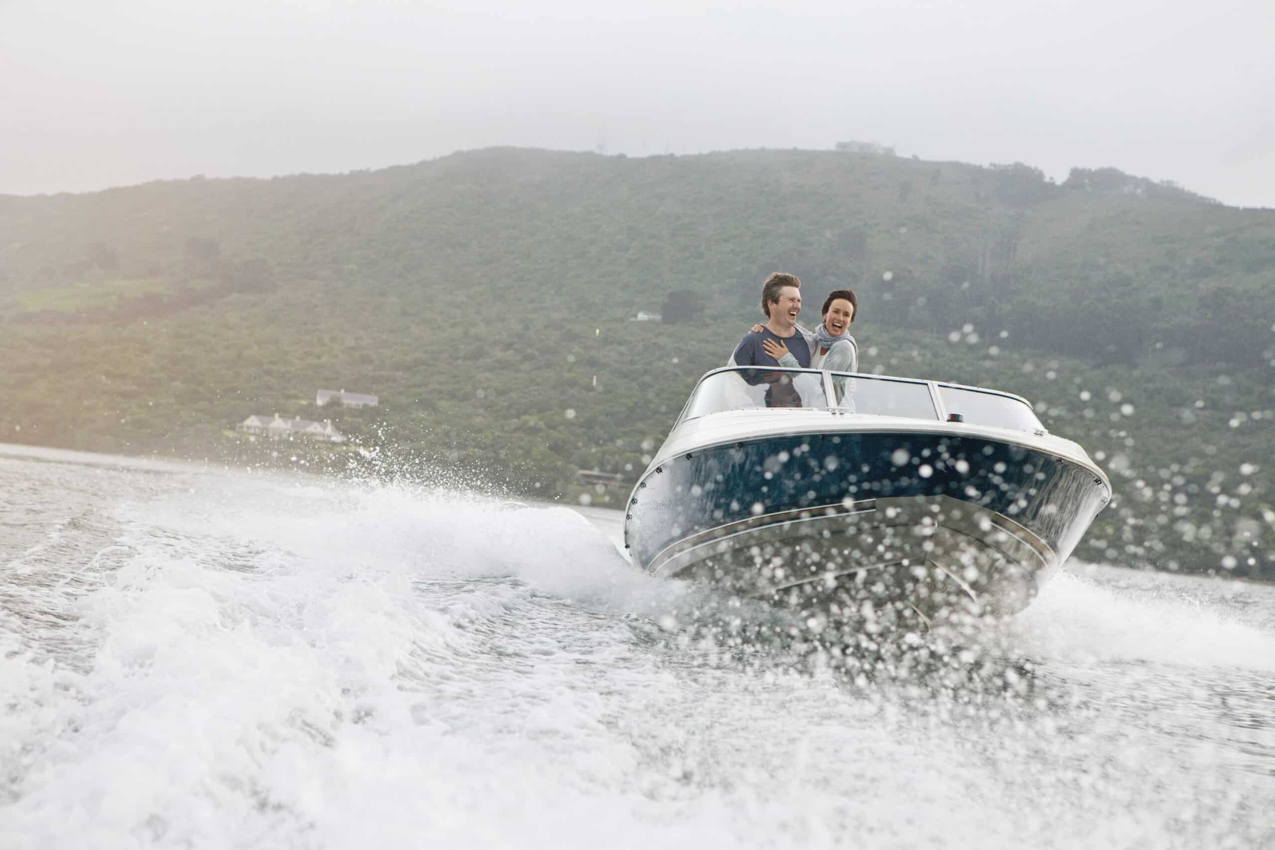 Zwei Personen genießen eine rasante Fahrt auf einem Motorboot, das im Wasser für Spritzer sorgt. Sie befinden sich auf einem See oder Fluss mit grünem, hügeligem Gelände im Hintergrund unter einem bewölkten Himmel. Beide Personen lächeln und scheinen Spaß zu haben. © Fotografie Tomas Rodriguez