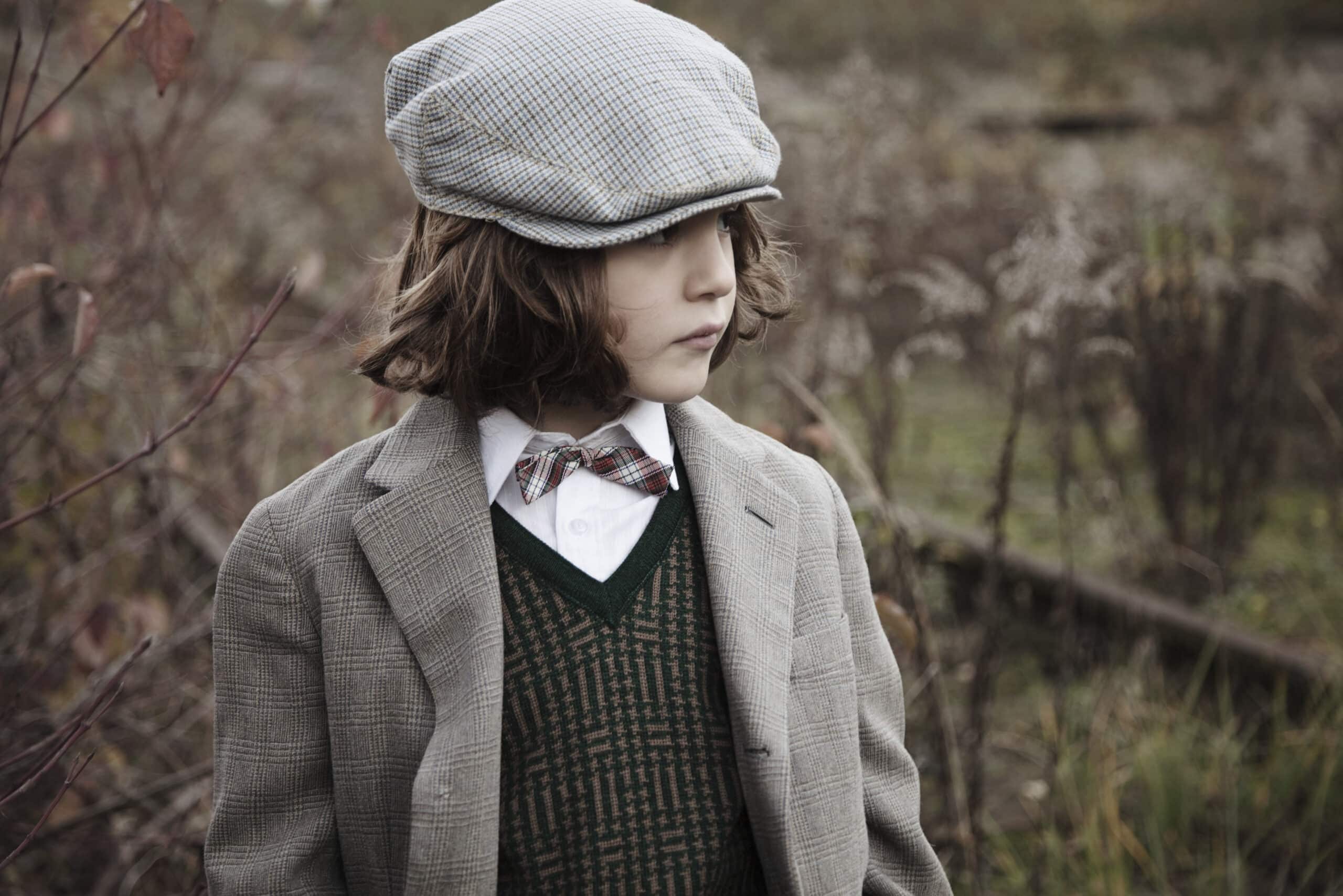 Ein kleines Kind mit schulterlangem braunem Haar trägt eine graue Schiebermütze, eine graue Tweedjacke, ein weißes Hemd, eine grüne Pullunderweste und eine karierte Fliege. Sie stehen draußen auf einem Feld mit überwucherter Vegetation und schauen zur Seite. © Fotografie Tomas Rodriguez