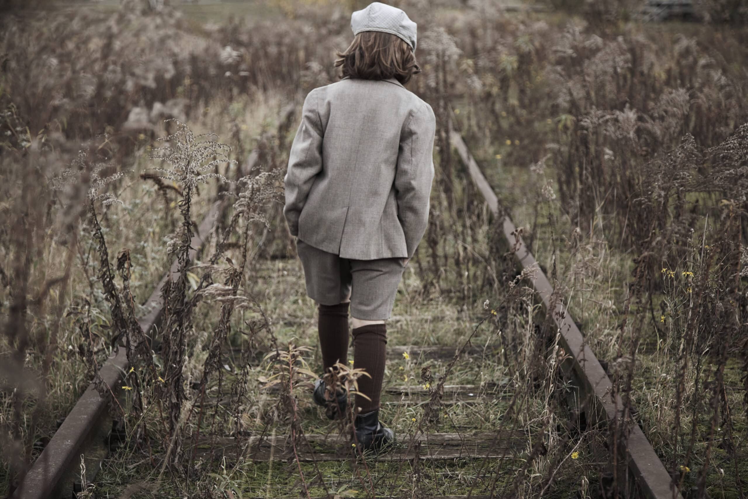 Ein kleines Kind mit schulterlangem Haar, einem grauen Anzug, Kniestrümpfen und einer Schiebermütze läuft an einem überwucherten und verlassenen Bahngleis entlang. Die umgebende Vegetation ist trocken und braun, was auf den beginnenden Herbst hindeutet. © Fotografie Tomas Rodriguez