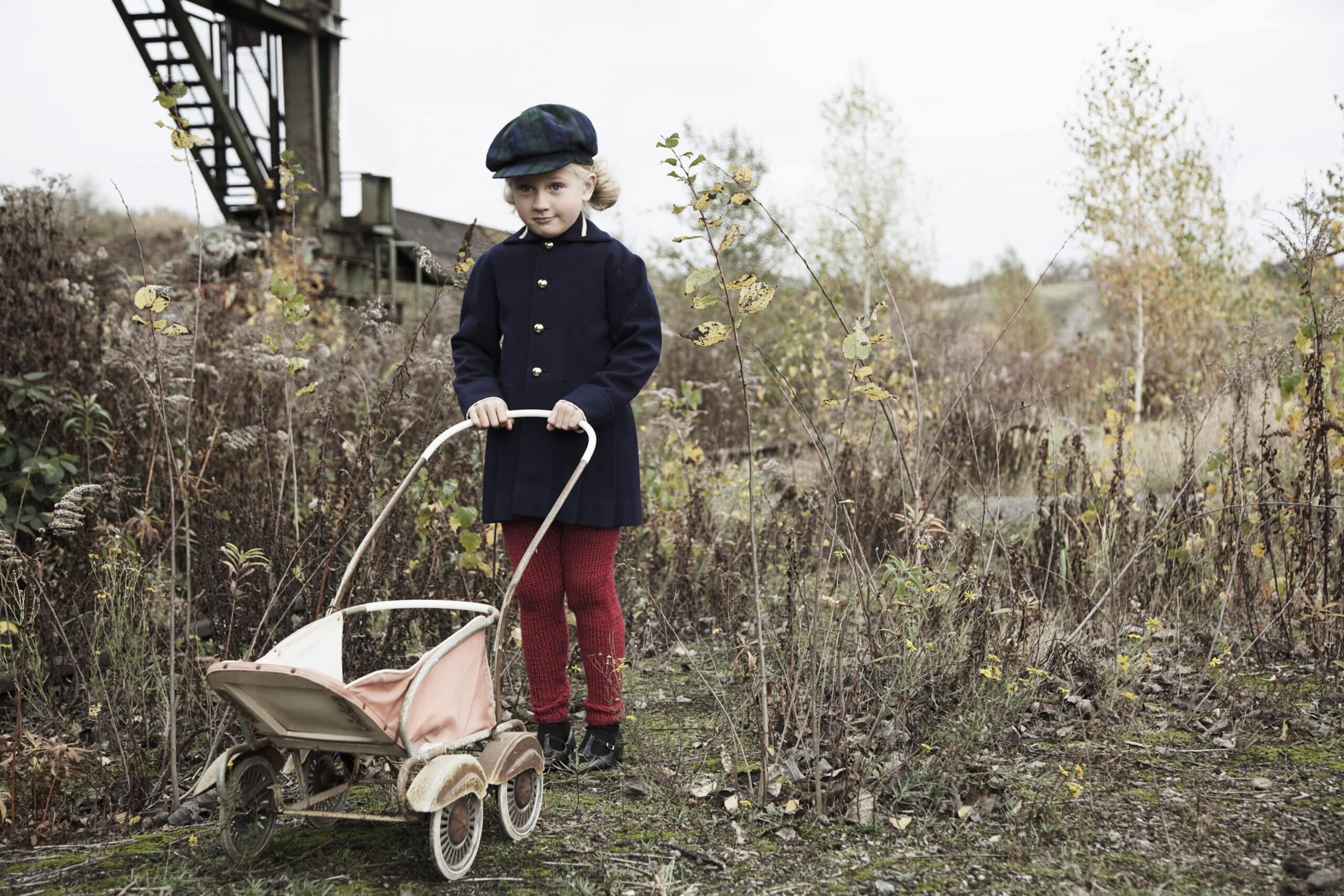 Ein Kind mit dunklem Mantel, Baskenmütze und roten Hosen steht auf einem verwilderten Feld und schiebt einen Spielzeug-Kinderwagen im Vintage-Stil. Im Hintergrund sind eine Industrieanlage und kahle Bäume unter einem bewölkten Himmel zu sehen. © Fotografie Tomas Rodriguez