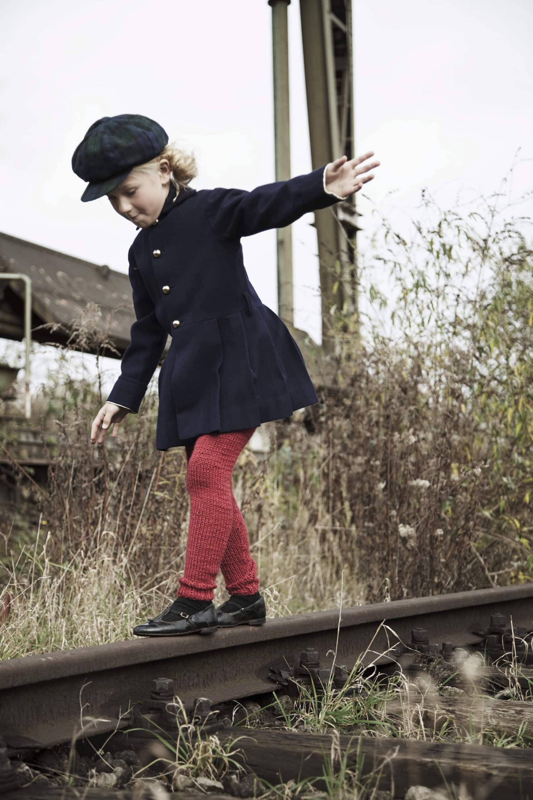 Ein Kind in einem marineblauen Mantel, roten Leggings, schwarzen Schuhen und einer schwarzen Baskenmütze läuft mit ausgestreckten Armen an einem Bahngleis entlang. Im Hintergrund sind überwuchertes Gras und ein Industriegebäude zu sehen. © Fotografie Tomas Rodriguez
