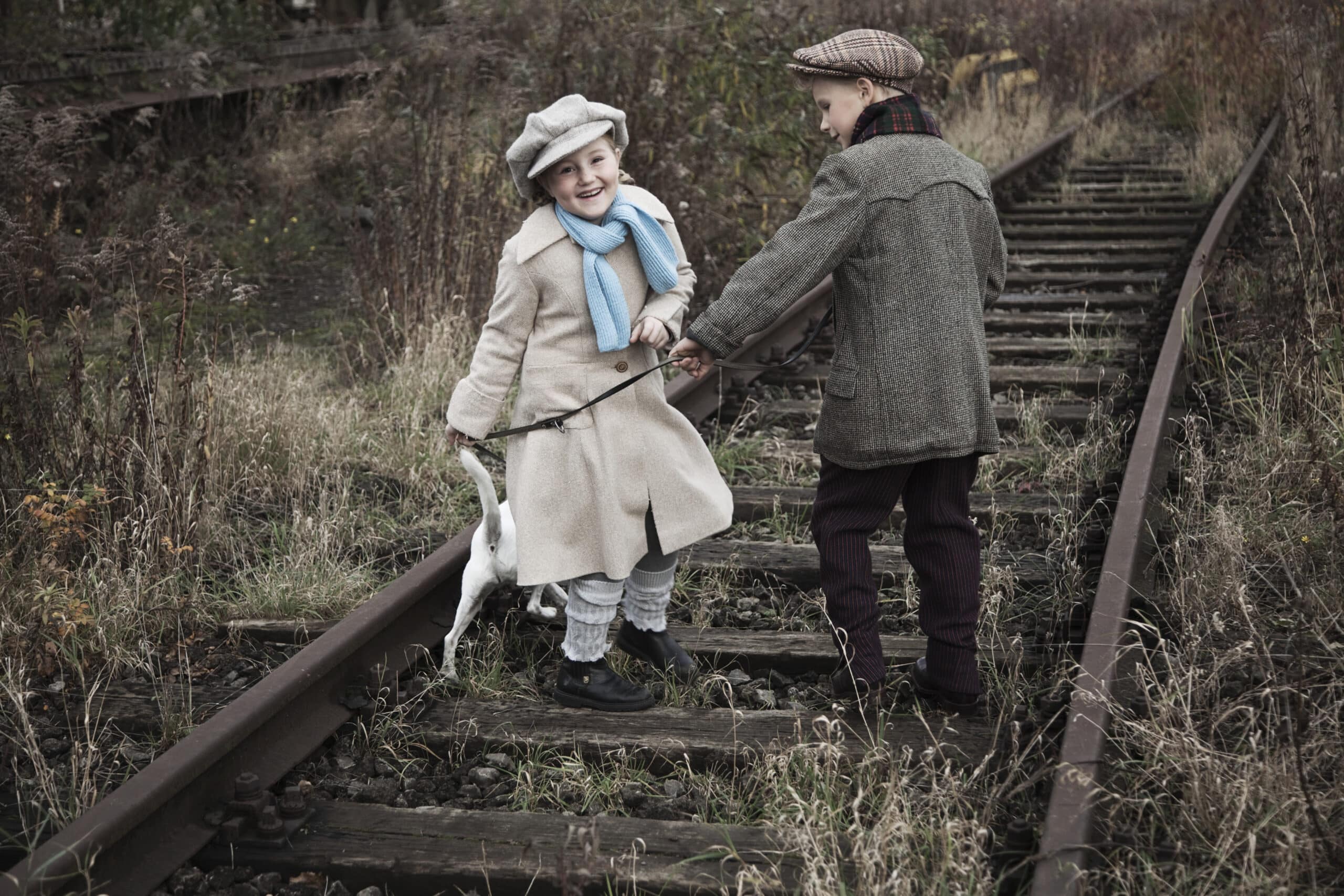 Zwei Kinder in Vintage-Kleidung, darunter Mützen und Schals, stehen auf alten, überwucherten Bahngleisen. Ein Kind lächelt und hält dabei einen Stock, an dem ein Spielzeugvogel baumelt, und das andere Kind schaut mit fröhlichem Gesichtsausdruck zu. © Fotografie Tomas Rodriguez