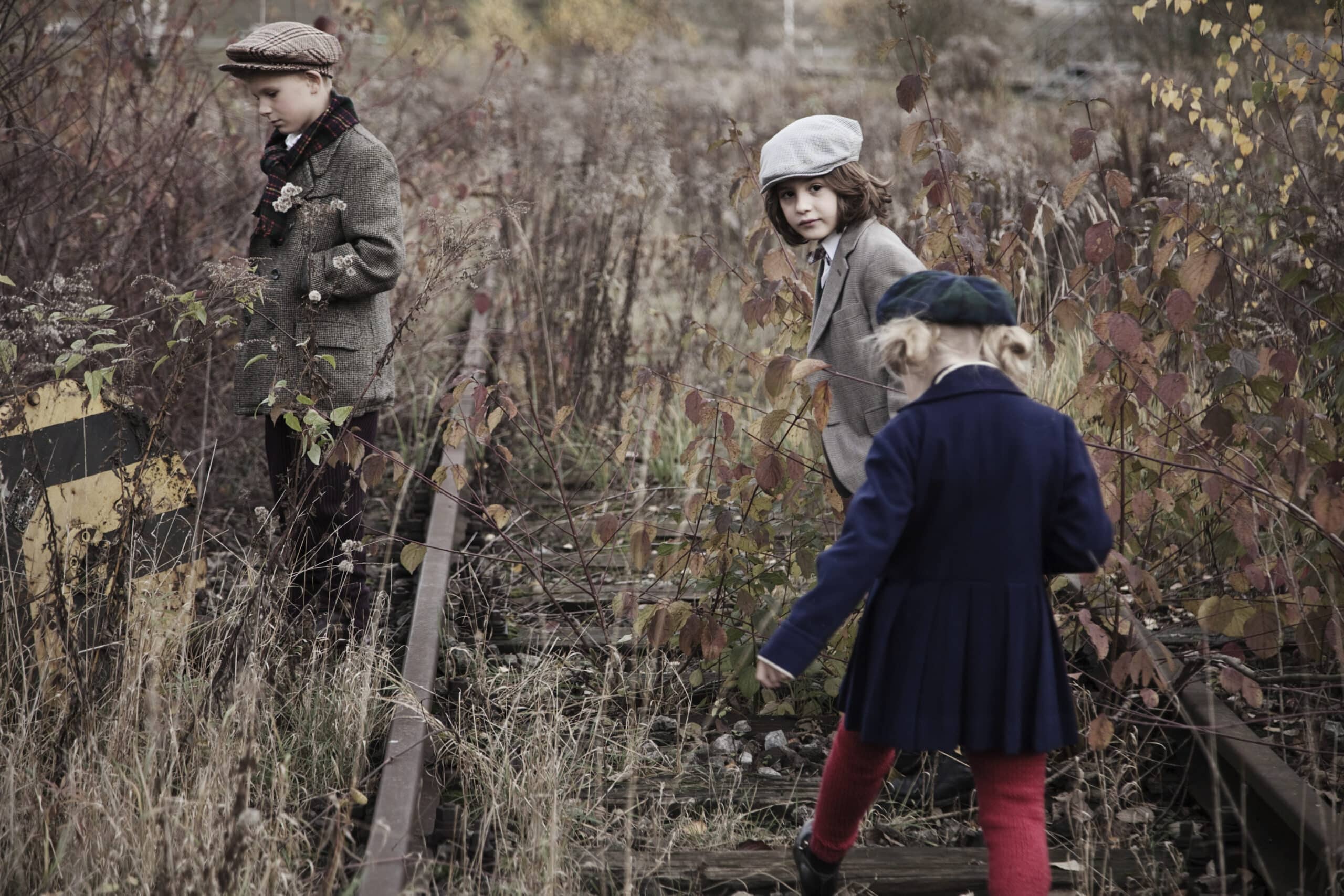 Drei Kinder in Vintage-Kleidung erkunden eine überwucherte und verlassene Bahnstrecke. Die Szene ist von herbstlichem Laub umgeben. Ein Kind blickt zurück, während die anderen beiden sich mit ihrer Umgebung beschäftigen und so eine nostalgische und abenteuerliche Atmosphäre schaffen. © Fotografie Tomas Rodriguez