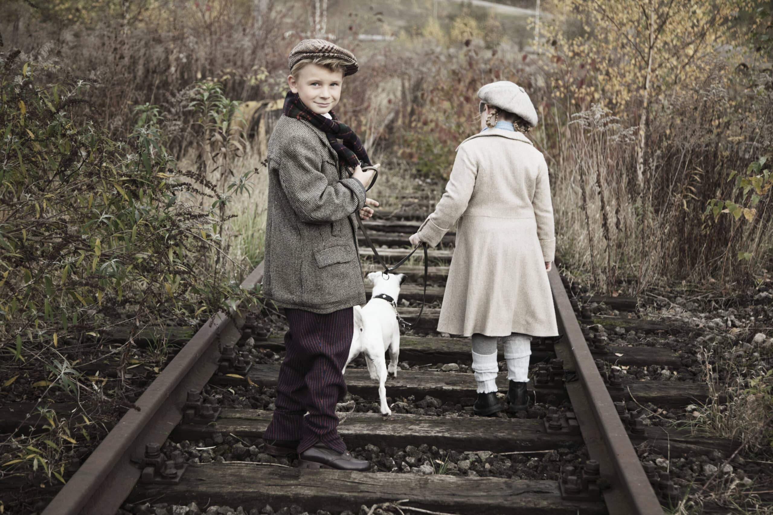 Ein Junge und ein Mädchen, beide in Vintage-Kleidung gekleidet, gehen mit einem weißen Hund an einem verlassenen Bahngleis entlang. Der Junge, der die Hundeleine hält, blickt zurück, während das Mädchen vorangeht. Die Umgebung ist mit herbstlichem Laub bewachsen. © Fotografie Tomas Rodriguez