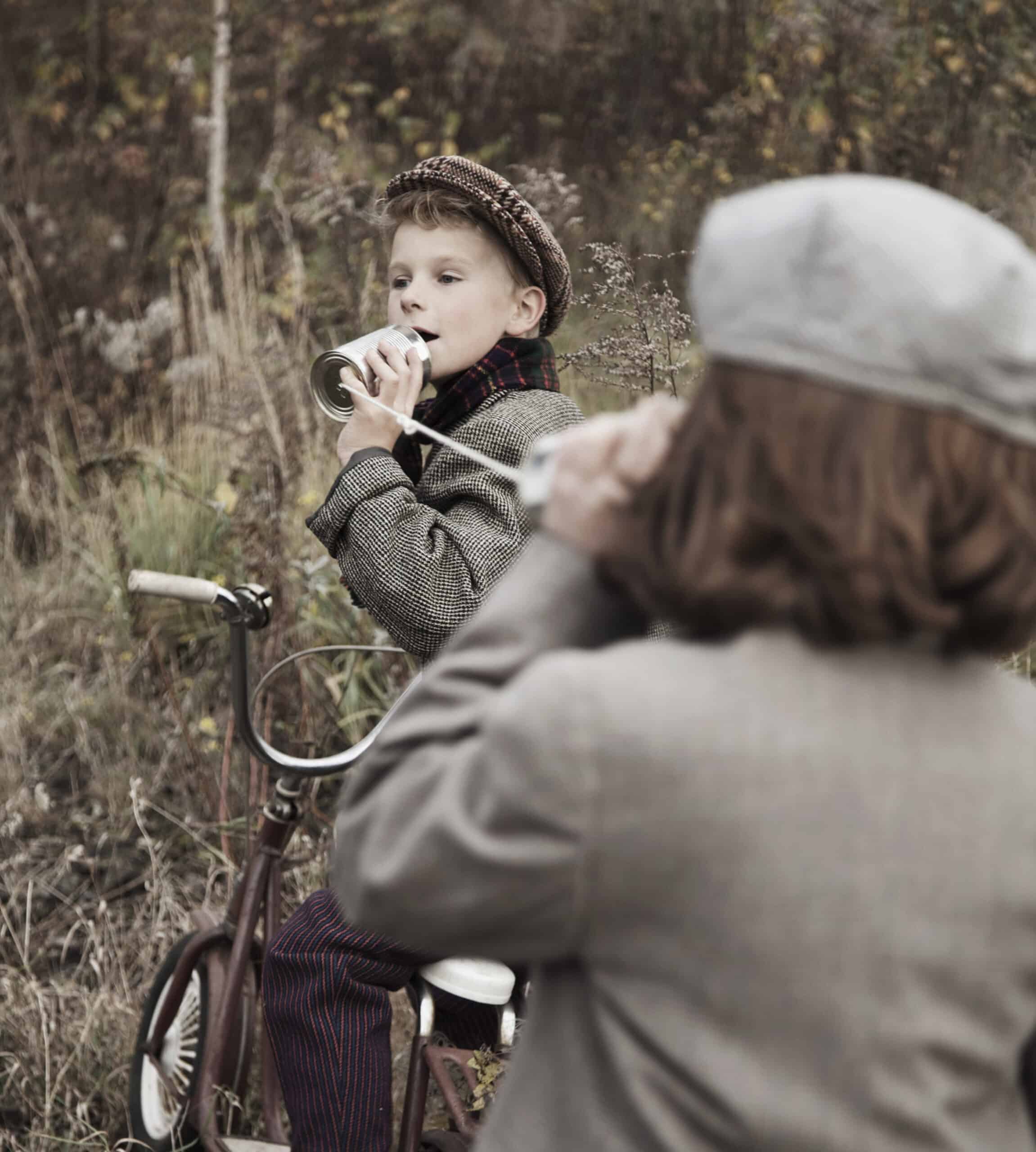 Zwei Kinder in Vintage-Kleidung spielen mit einem Dosentelefon. Ein Kind sitzt auf einem Dreirad und hält eine Dose ans Ohr, während das andere Kind in eine Dose spricht, die mit einer Schnur verbunden ist. Sie befinden sich draußen in einem grasbewachsenen Waldgebiet. © Fotografie Tomas Rodriguez