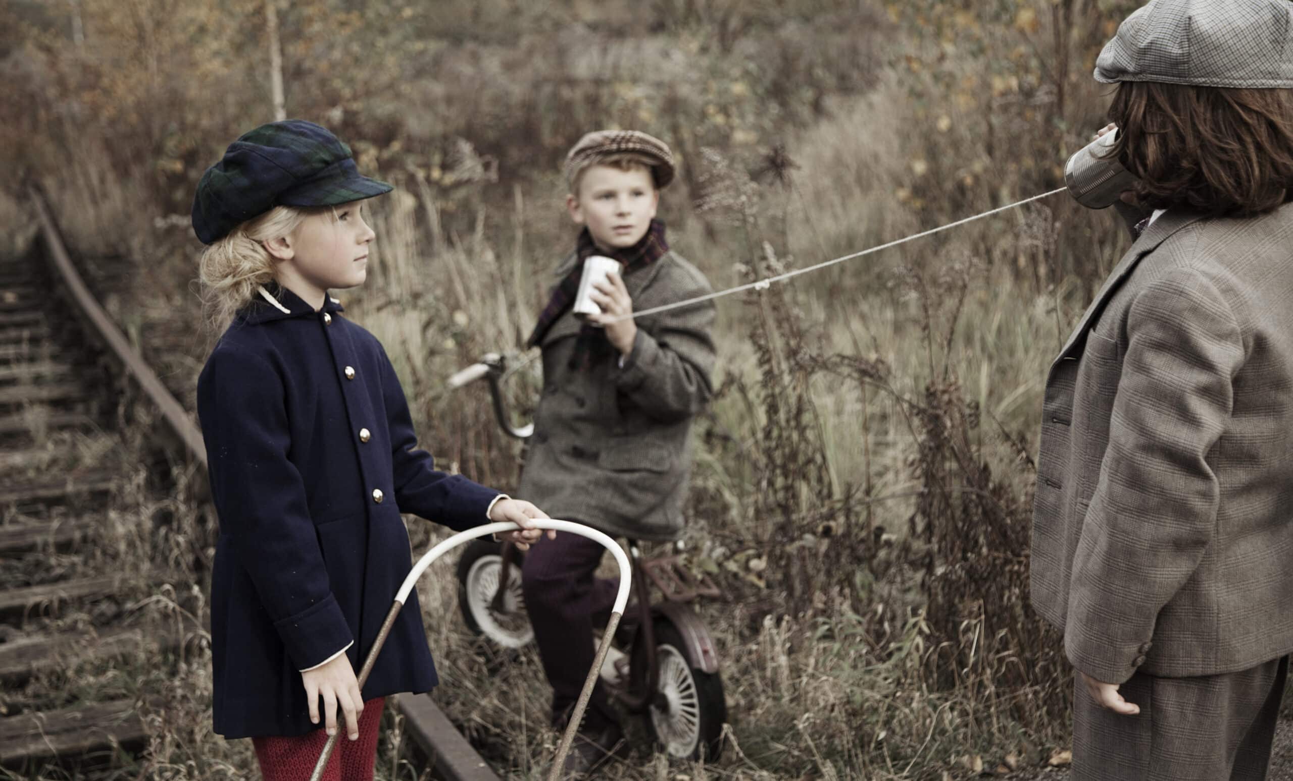 Drei Kinder in Vintage-Kleidung spielen neben alten, unbenutzten Bahngleisen. Ein Kind, das einen blauen Mantel und eine grüne Mütze trägt, hält einen weißen Reifen. Ein anderes Kind, das einen grauen Mantel und eine Schiebermütze trägt, sitzt auf einem Dreirad und spricht in eine Blechdose, die durch eine Schnur mit einem dritten Kind verbunden ist, das zuhört. © Fotografie Tomas Rodriguez