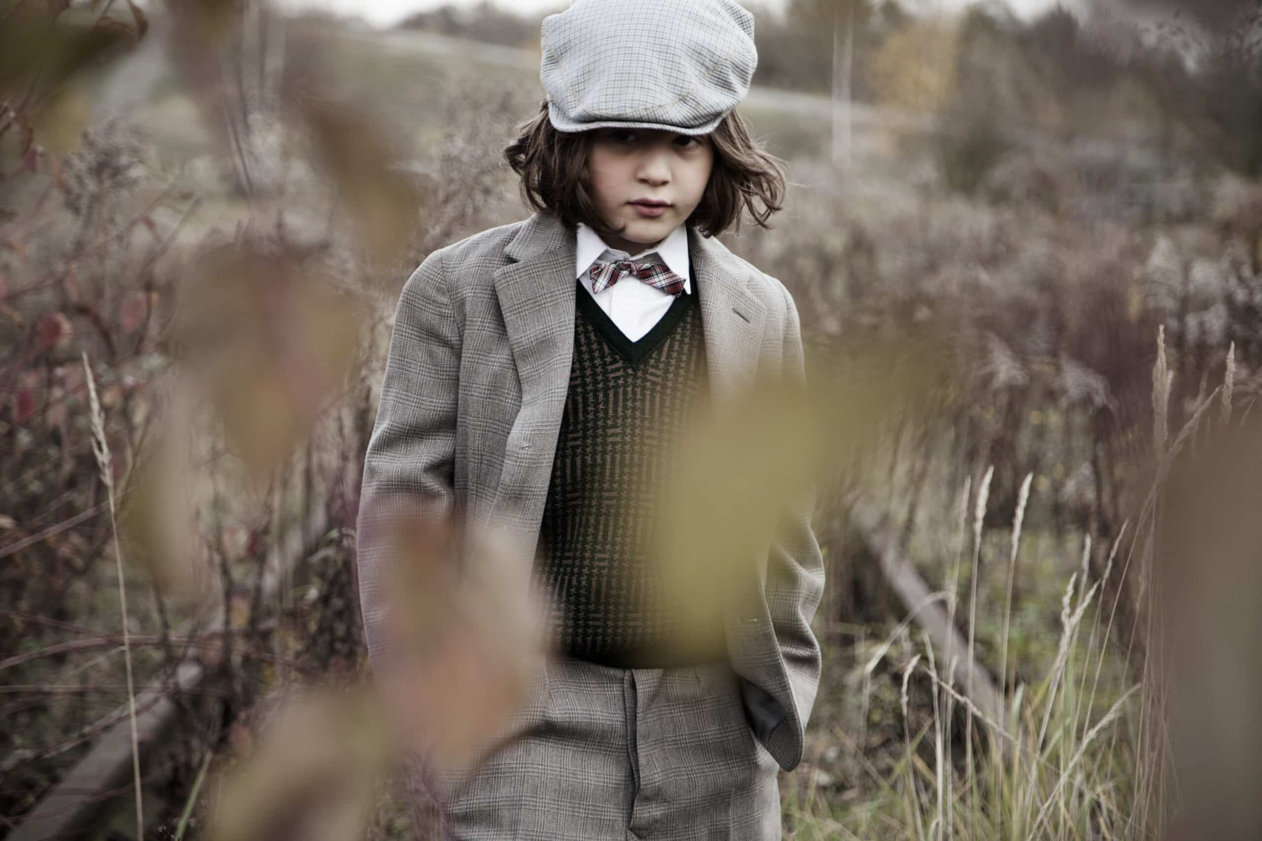 Ein kleiner Junge in einem Vintage-Outfit, darunter eine karierte Schiebermütze, ein karierter Blazer und eine Fliege, steht draußen auf einem Feld mit wildem Gras und Pflanzen. Die Szene ist rustikal und nostalgisch, mit sanften, gedämpften Farben. Der Junge sieht nachdenklich und ernst aus. © Fotografie Tomas Rodriguez