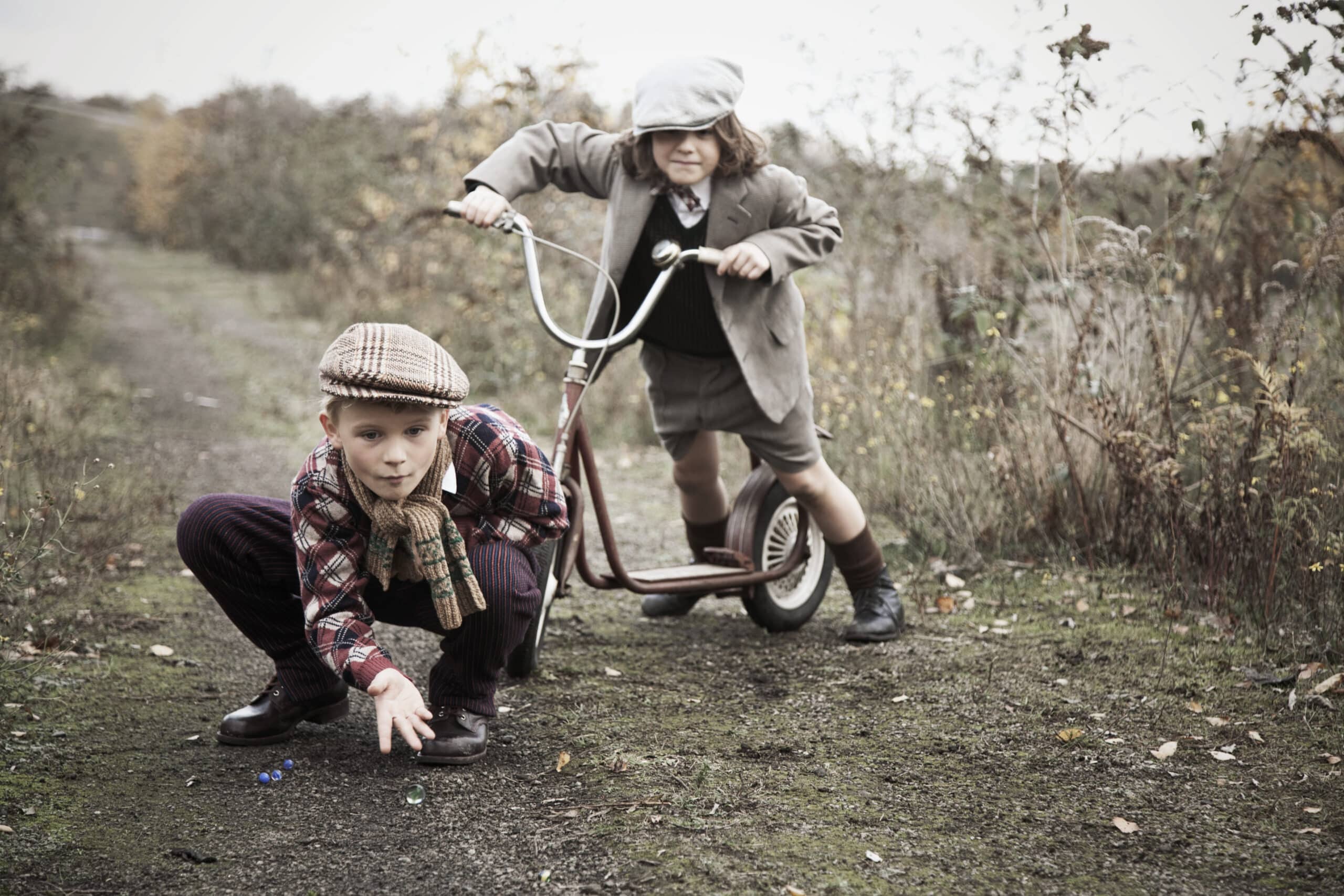 Zwei junge Jungen in Vintage-Kleidung spielen auf einem Feldweg, der von Gras und Sträuchern umgeben ist. Ein Junge hockt auf dem Boden und spielt mit Murmeln. Der andere steht auf einem klassischen Tretroller und beobachtet ihn aufmerksam. Beide tragen Mützen und wirken, als würden sie mit ihren Aktivitäten beschäftigt sein. © Fotografie Tomas Rodriguez