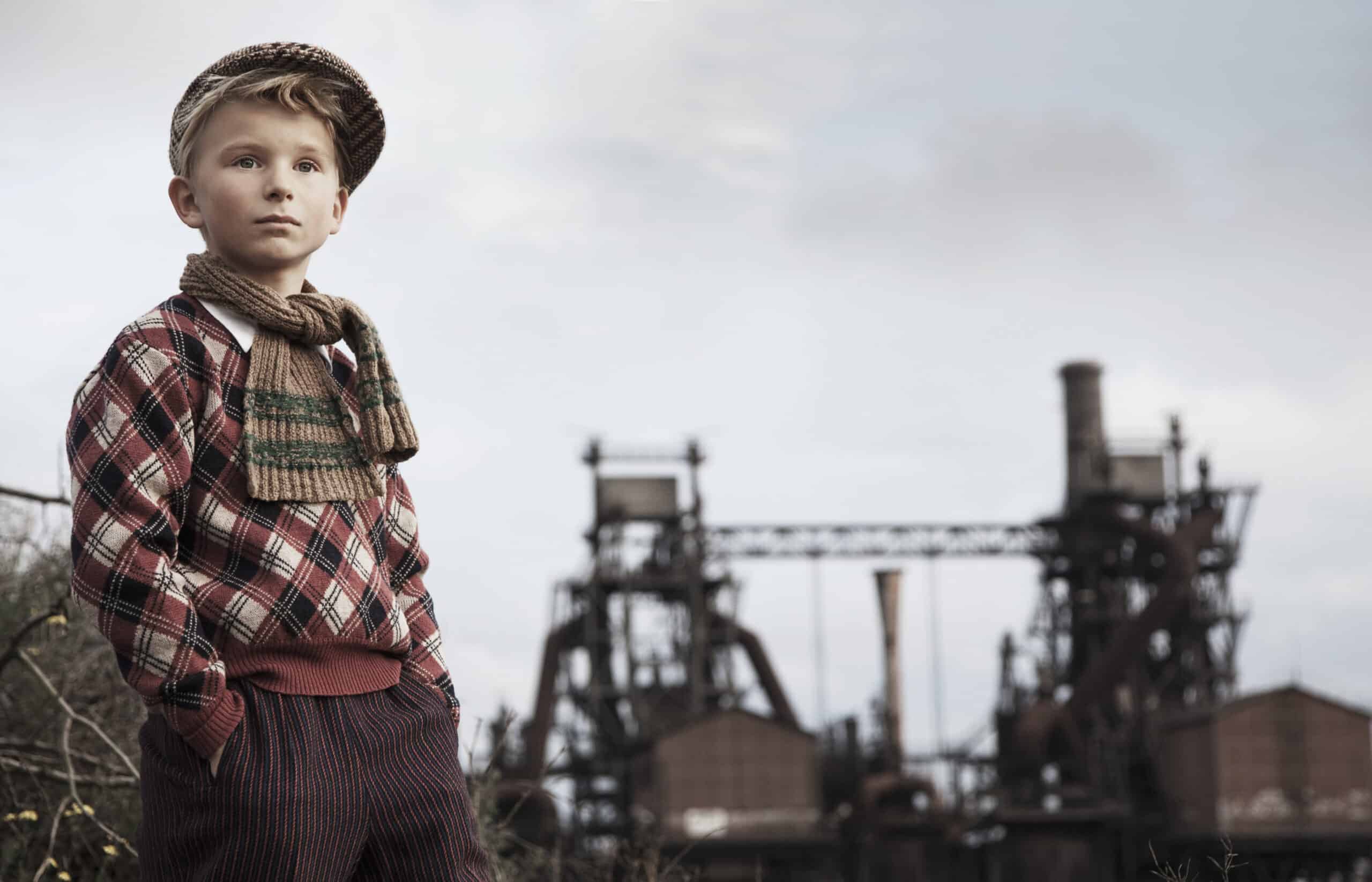 Ein kleiner Junge in Vintage-Kleidung steht mit den Händen in den Taschen da und blickt zur Seite. Er trägt ein kariertes Hemd, gestreifte Hosen, einen Schal und eine Zeitungsjungenmütze. Im Hintergrund sind Industrieanlagen vor einem bewölkten Himmel zu sehen. © Fotografie Tomas Rodriguez