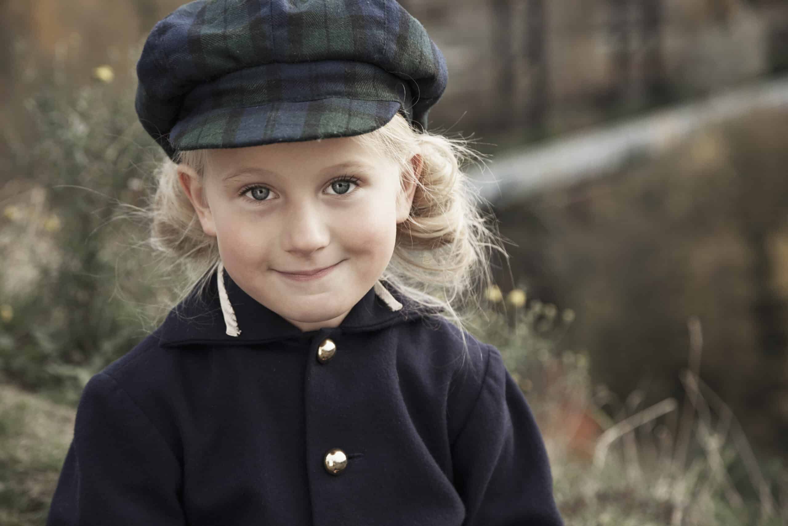 Ein kleines Kind mit hellem Haar und Zöpfen lächelt in die Kamera. Es trägt einen dunklen Mantel mit goldenen Knöpfen und eine dunkelgrün karierte Schiebermütze. Der Hintergrund ist verschwommen und zeigt eine natürliche Umgebung im Freien mit viel Grün. © Fotografie Tomas Rodriguez