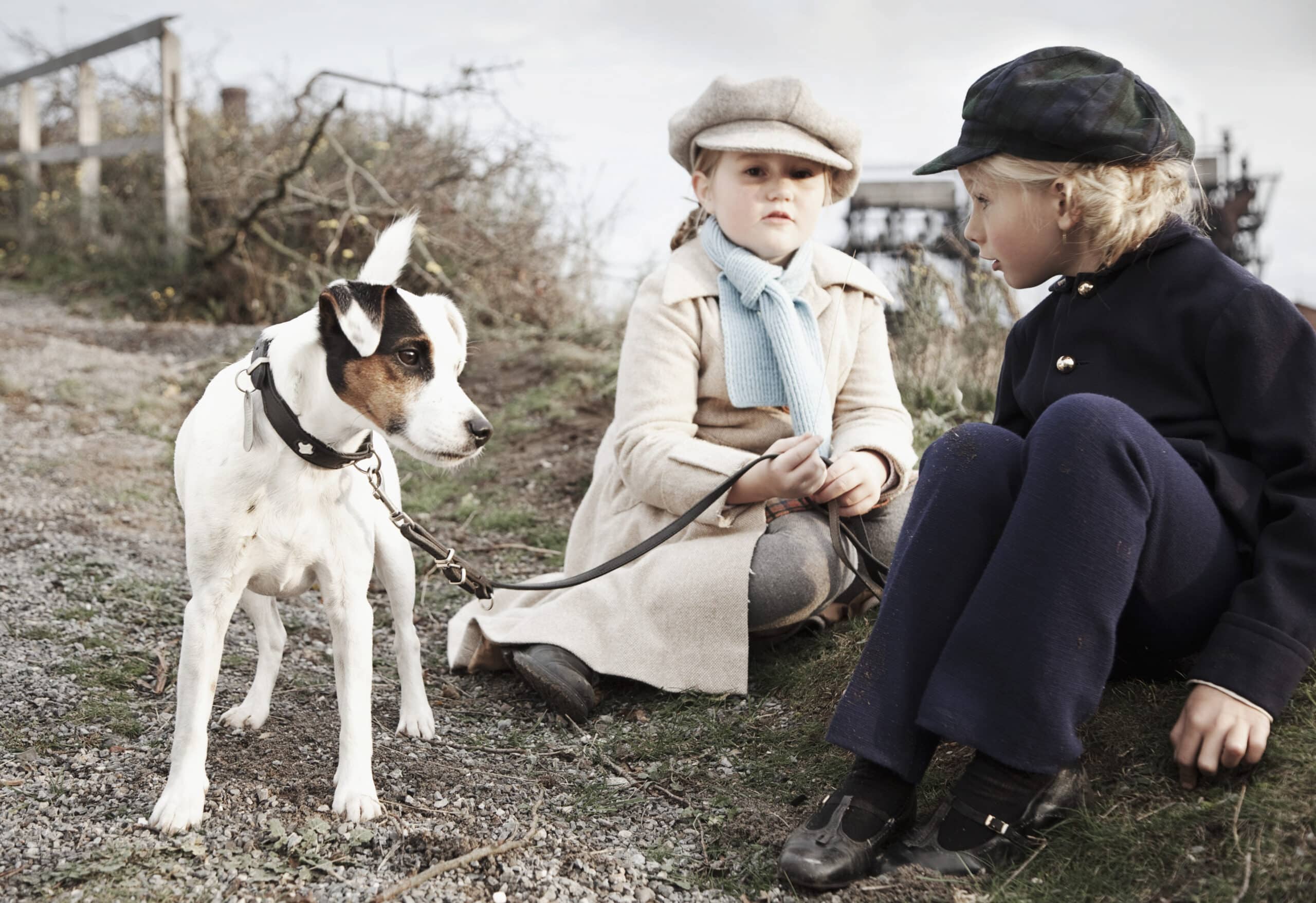 Zwei Kinder in Vintage-Kleidung sitzen auf einem Grasweg. Das Kind links hält die Leine eines kleinen schwarz-weißen Hundes, der in der Nähe steht. Das Kind rechts sitzt mit den Händen auf dem Boden und schaut den Hund an. Im Hintergrund ist ein bewölkter Himmel zu sehen. © Fotografie Tomas Rodriguez