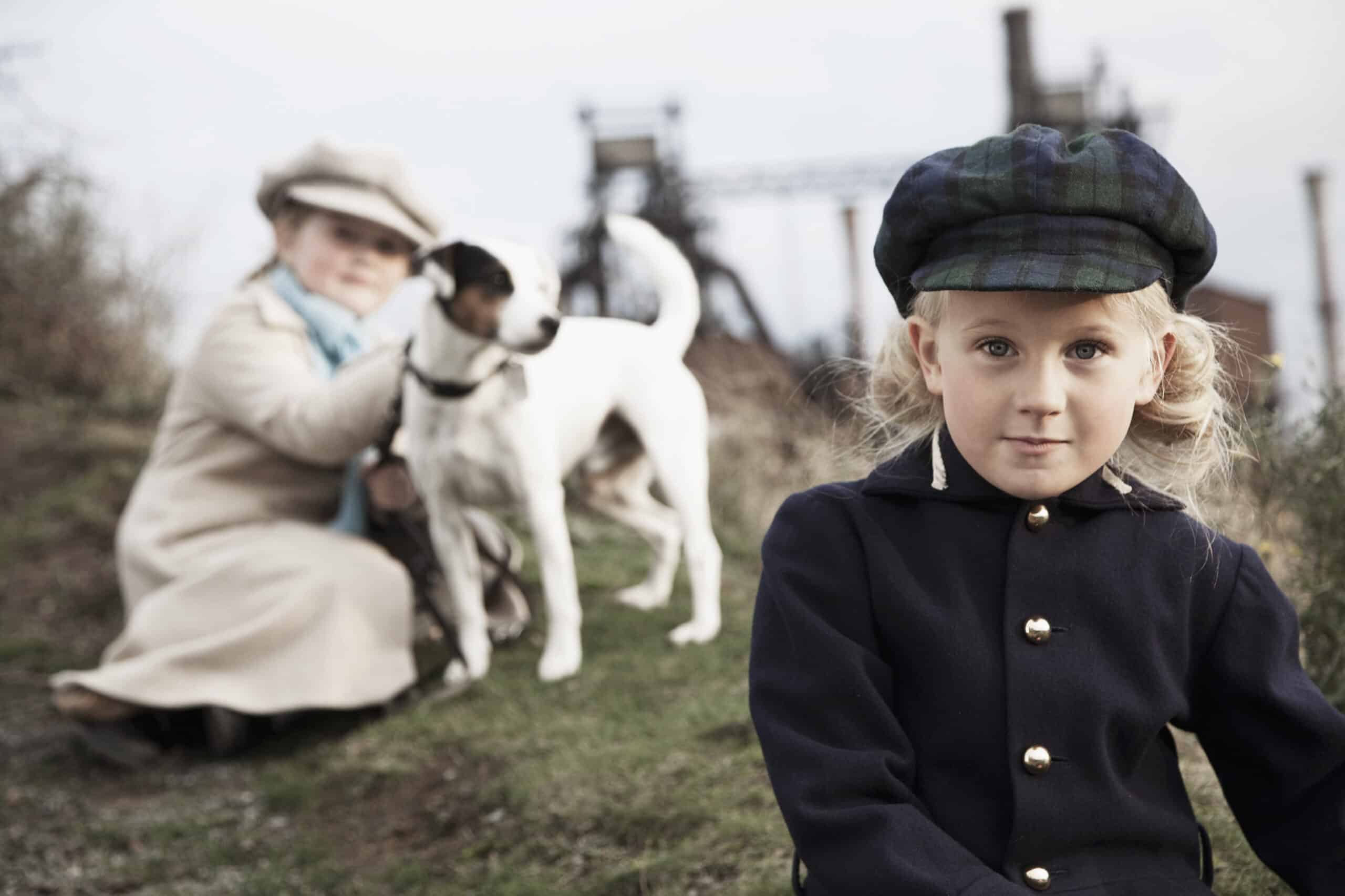 Zwei Kinder und ein kleiner Hund sind draußen. Ein Kind, in marineblauem Mantel und Mütze, sitzt im Vordergrund und schaut in die Kamera. Das andere Kind, in helle Kleidung und Hut gekleidet, ist im Hintergrund und hält den Hund. In der Ferne ist ein Industriegebäude zu sehen. © Fotografie Tomas Rodriguez