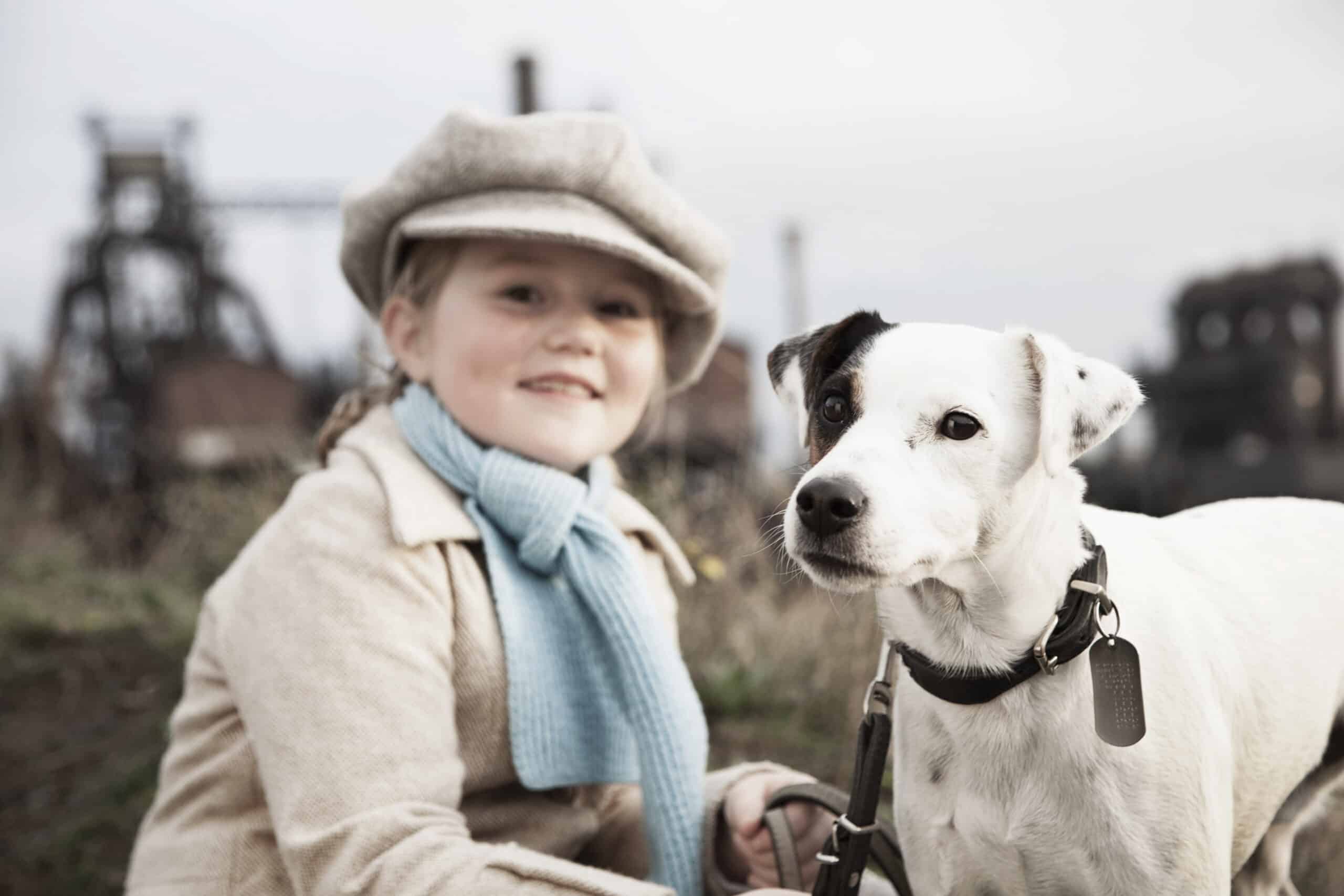 Ein kleines Kind mit beigem Mantel, hellblauem Schal und Schiebermütze sitzt lächelnd neben einem weißen Hund mit schwarzen Flecken an den Ohren. Der Hund trägt ein Halsband mit Anhänger und eine Leine. Im Hintergrund ist eine industrielle Kulisse mit verschwommenen Strukturen zu sehen. © Fotografie Tomas Rodriguez