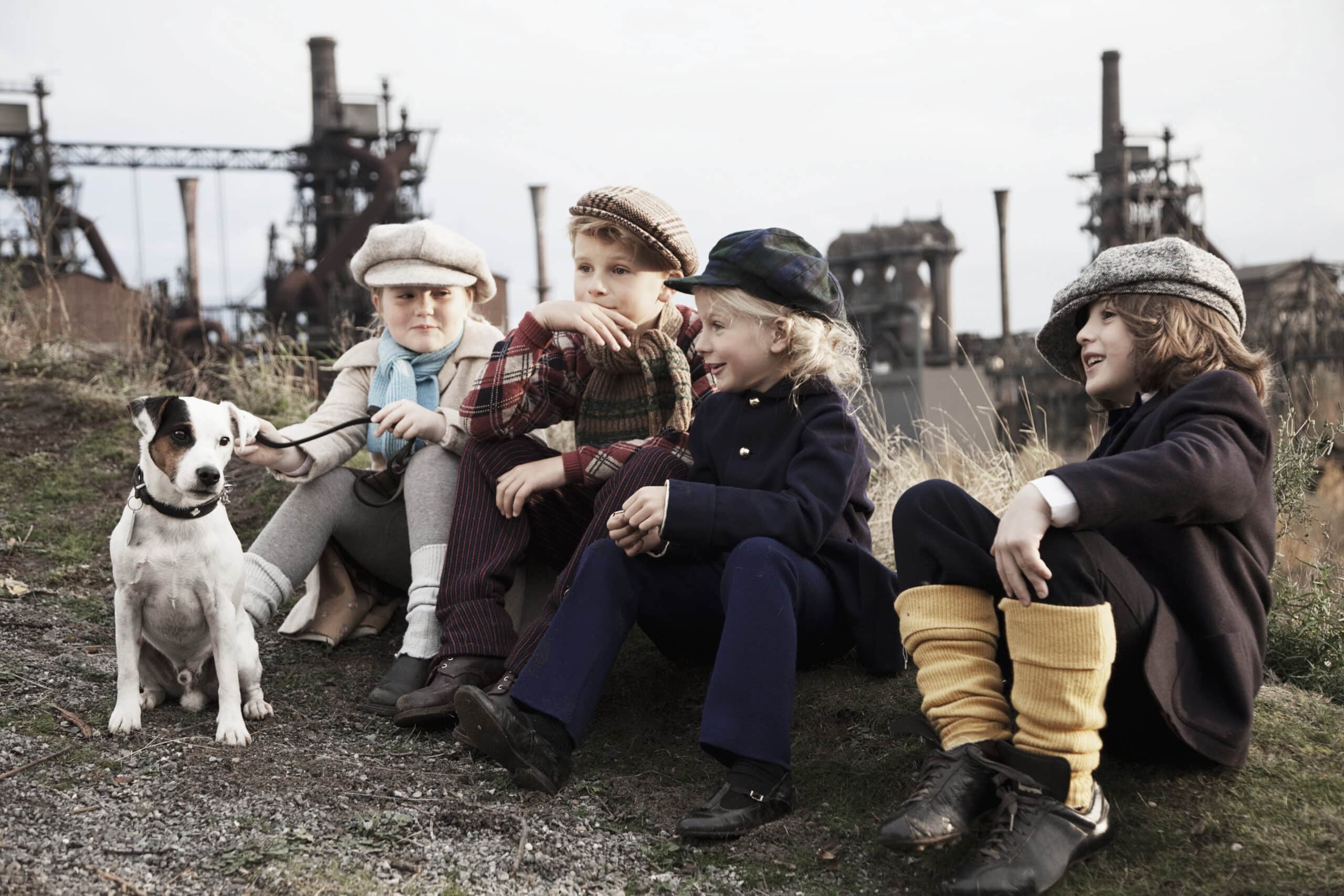 Vier Kinder in Vintage-Kleidung sitzen draußen auf dem Boden. Im Hintergrund sieht man große Fabriken und Schornsteine. Ein Kind hält die Leine eines kleinen Hundes, der in der Nähe sitzt. Die Gruppe wirkt fröhlich und entspannt. © Fotografie Tomas Rodriguez