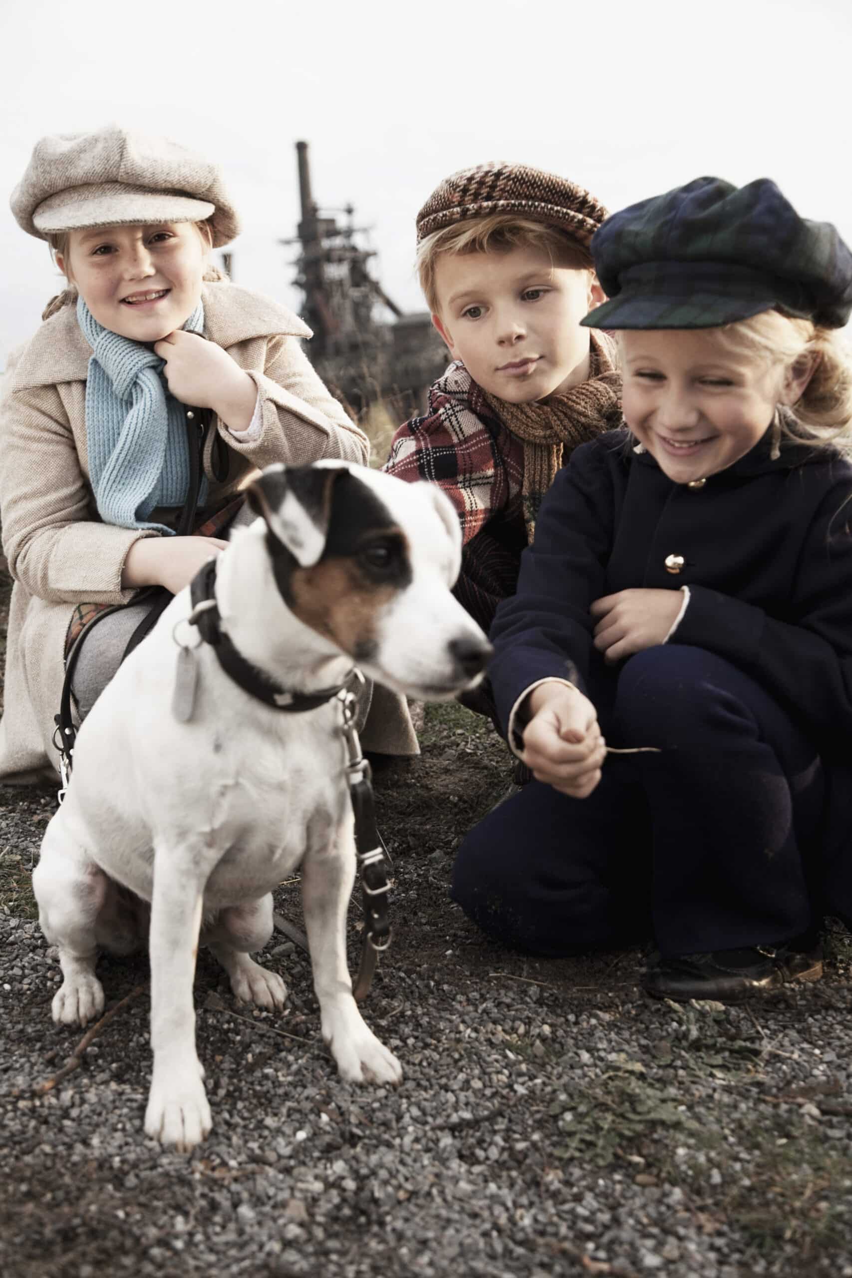 Drei Kinder, die Vintage-Kleidung wie Mützen, Mäntel und Schals tragen, sitzen dicht beieinander im Freien. Vor ihnen steht ein weißer Hund mit schwarzen und braunen Markierungen auf dem Kopf, der zur Seite schaut. Die Kinder scheinen glücklich und mit dem Hund beschäftigt zu sein. © Fotografie Tomas Rodriguez