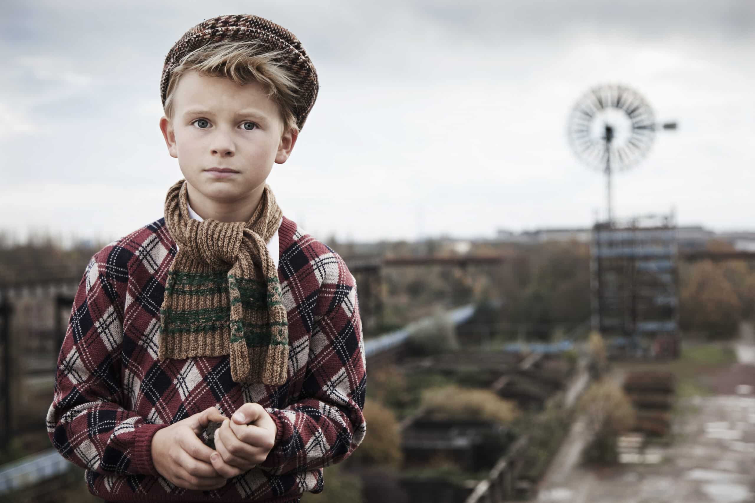 Ein kleiner Junge mit kariertem Hemd, gestreiftem Schal und Schiebermütze steht im Freien vor einem verschwommenen industriellen Hintergrund. Er hält einen kleinen Gegenstand in den Händen und blickt mit ernster Miene direkt in die Kamera. © Fotografie Tomas Rodriguez