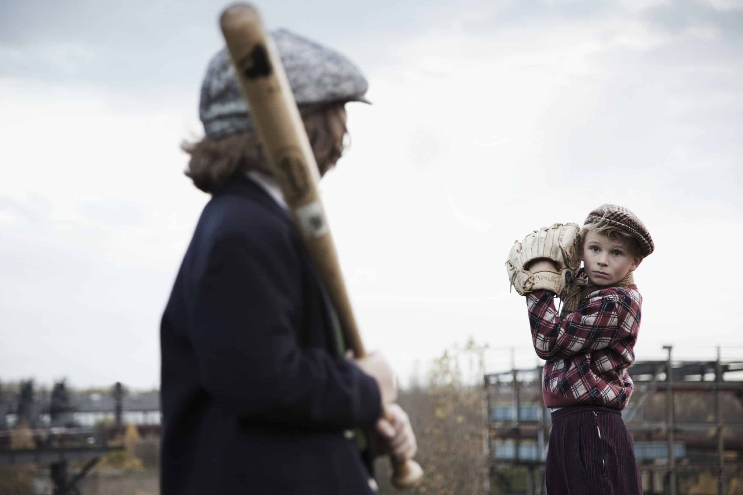 Zwei junge Jungen in Vintage-Kleidung spielen im Freien Baseball. Ein Junge hält einen Schläger und blickt von der Kamera weg, während der andere Junge, der eine Schiebermütze und ein kariertes Hemd trägt, sich mit erhobenem Handschuh darauf vorbereitet, einen Baseball zu werfen. Ein industrieller Hintergrund ist sichtbar. © Fotografie Tomas Rodriguez