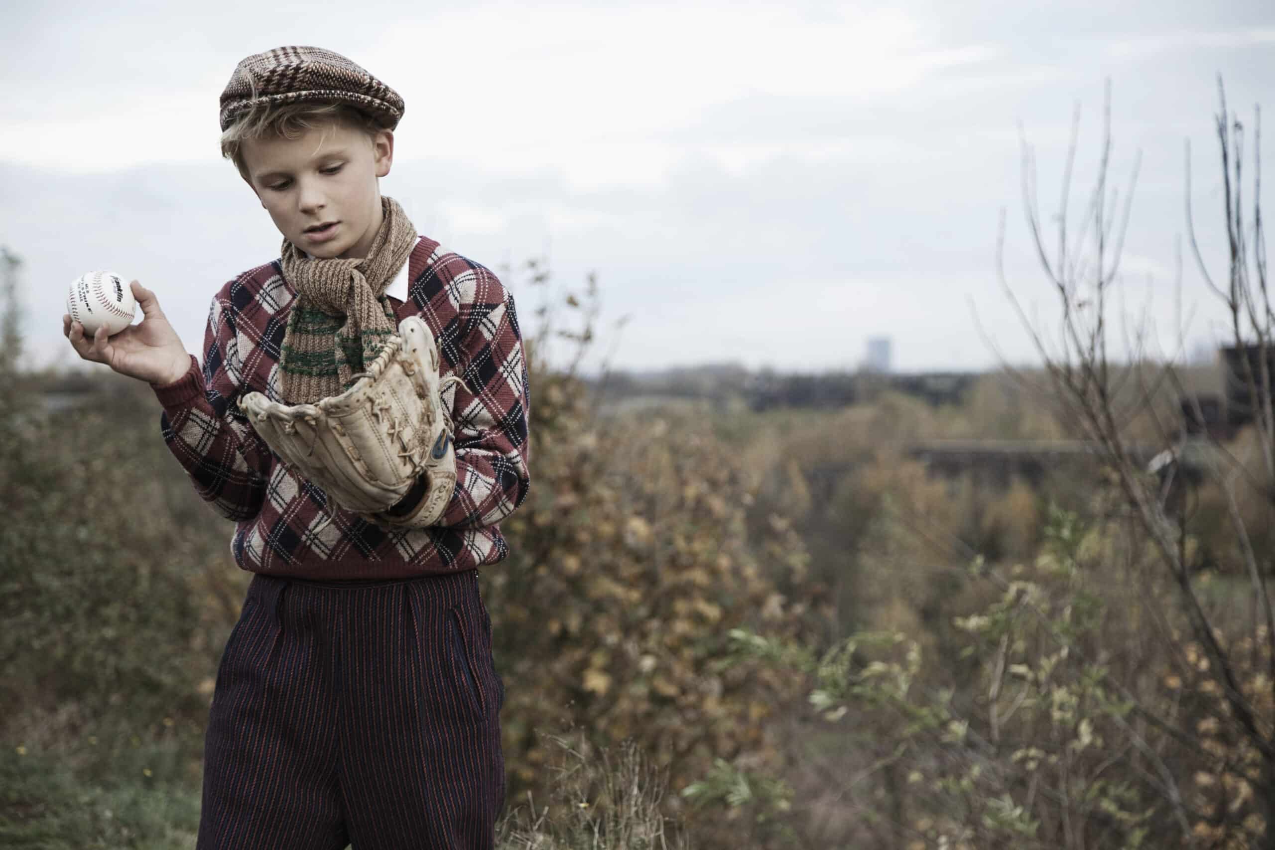 Ein kleiner Junge mit Schiebermütze, Schal und kariertem Hemd steht im Freien, hält einen Baseball und einen Handschuh in der Hand und blickt auf sie herab. Im Hintergrund ist eine verschwommene Naturlandschaft mit Bäumen und einem bewölkten Himmel zu sehen. © Fotografie Tomas Rodriguez