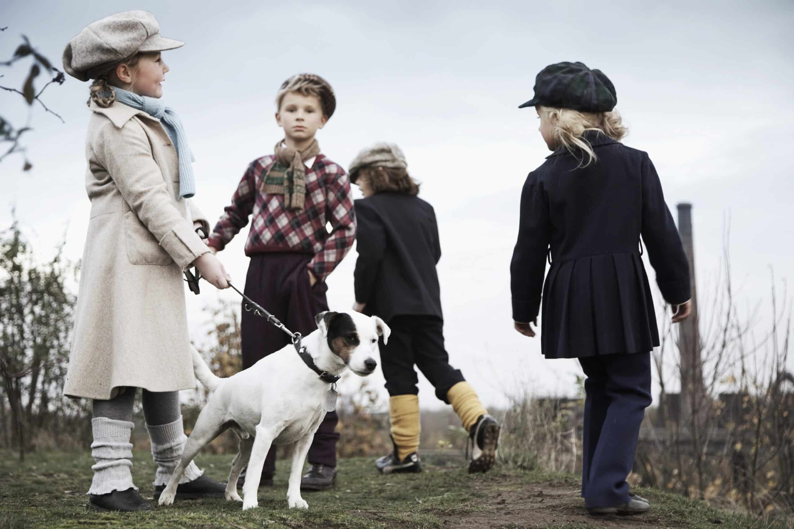 Vier Kinder sind an einem bewölkten Tag draußen. Ein Kind hält einen Hund an der Leine. Sie sind alle in Vintage-Kleidung gekleidet, darunter Mützen, Schals und Kniestrümpfe. Der Hintergrund ist ein bedeckter Himmel mit Bäumen und einer verschwommenen Industrieanlage. © Fotografie Tomas Rodriguez