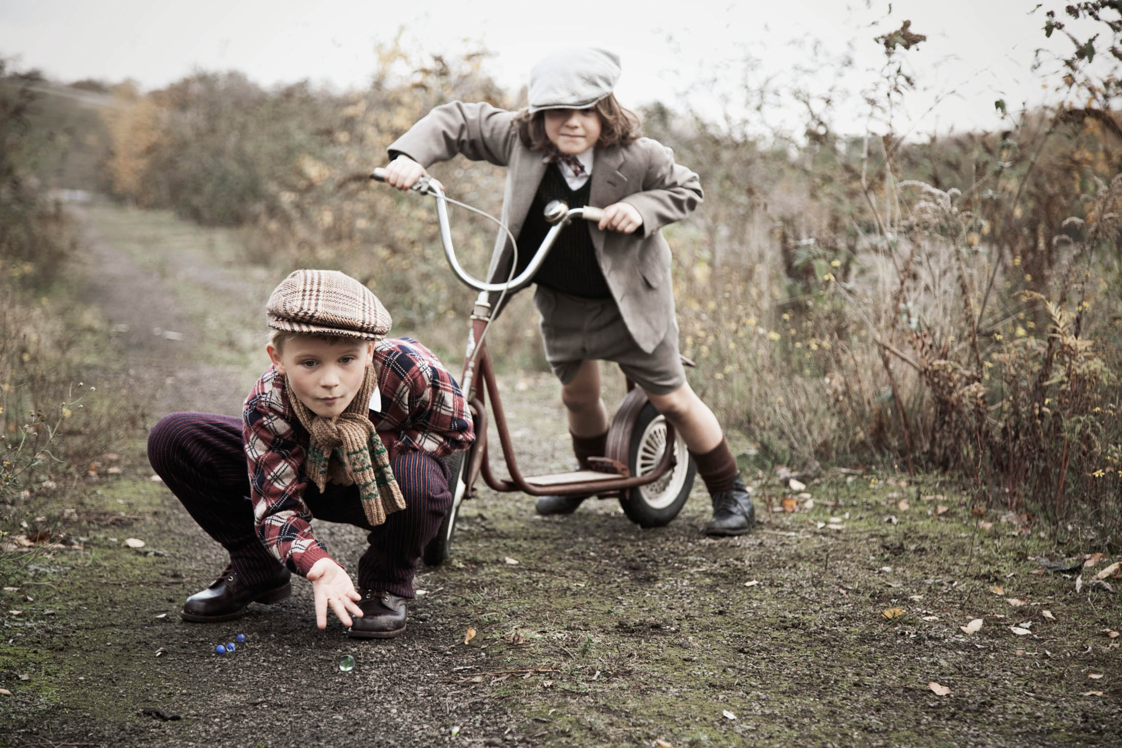 Zwei Kinder spielen im Freien; eines kniet und hebt Murmeln auf, während das andere mit einem Roller auf einem Feldweg steht. Beide tragen Vintage-Kleidung. © Fotografie Tomas Rodriguez