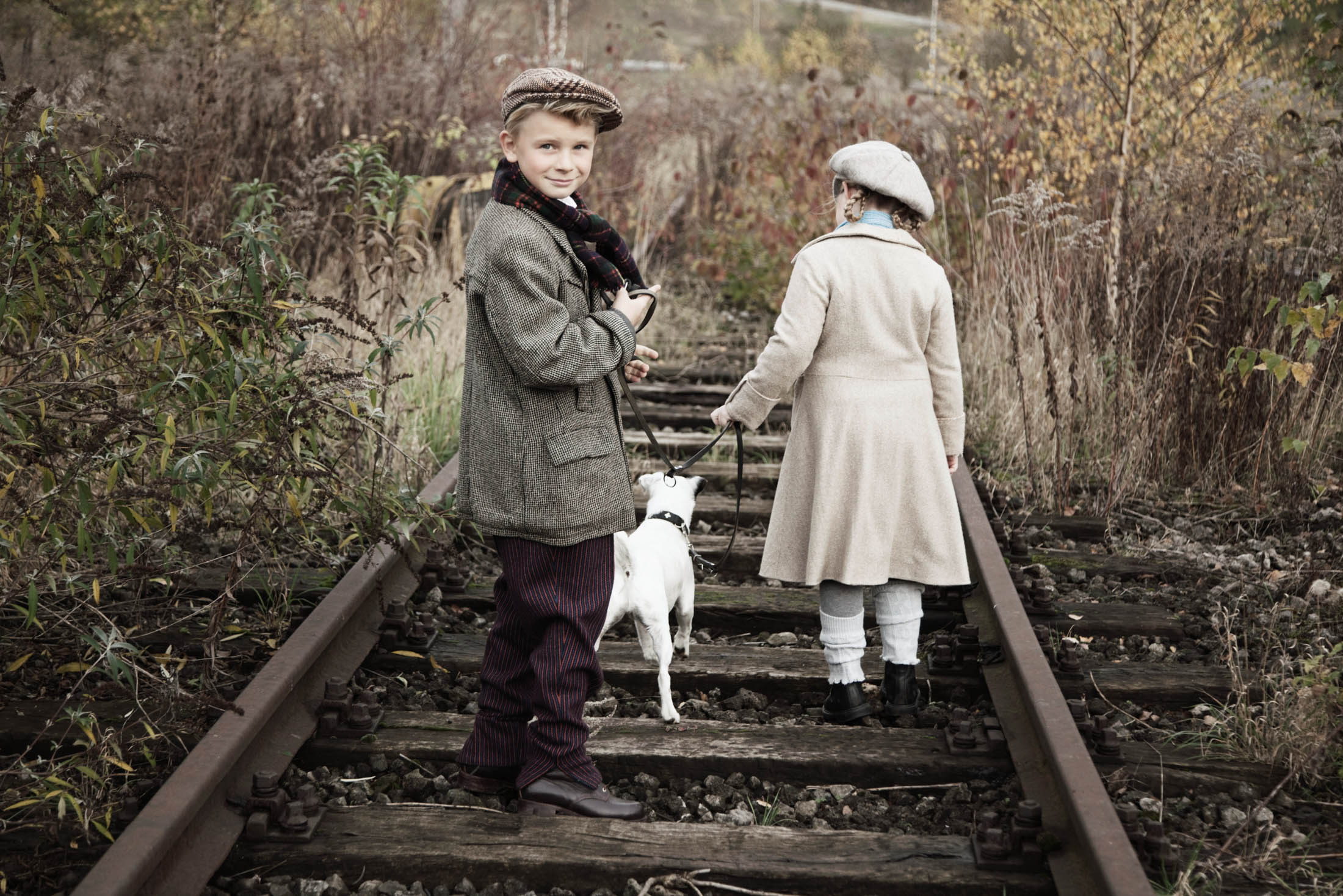 Zwei Kinder in Vintage-Kleidung gehen mit einem kleinen weißen Hund an stillgelegten Bahngleisen entlang. Der Junge blickt in die Kamera, während das Mädchen wegblickt. Herbststimmung. © Fotografie Tomas Rodriguez