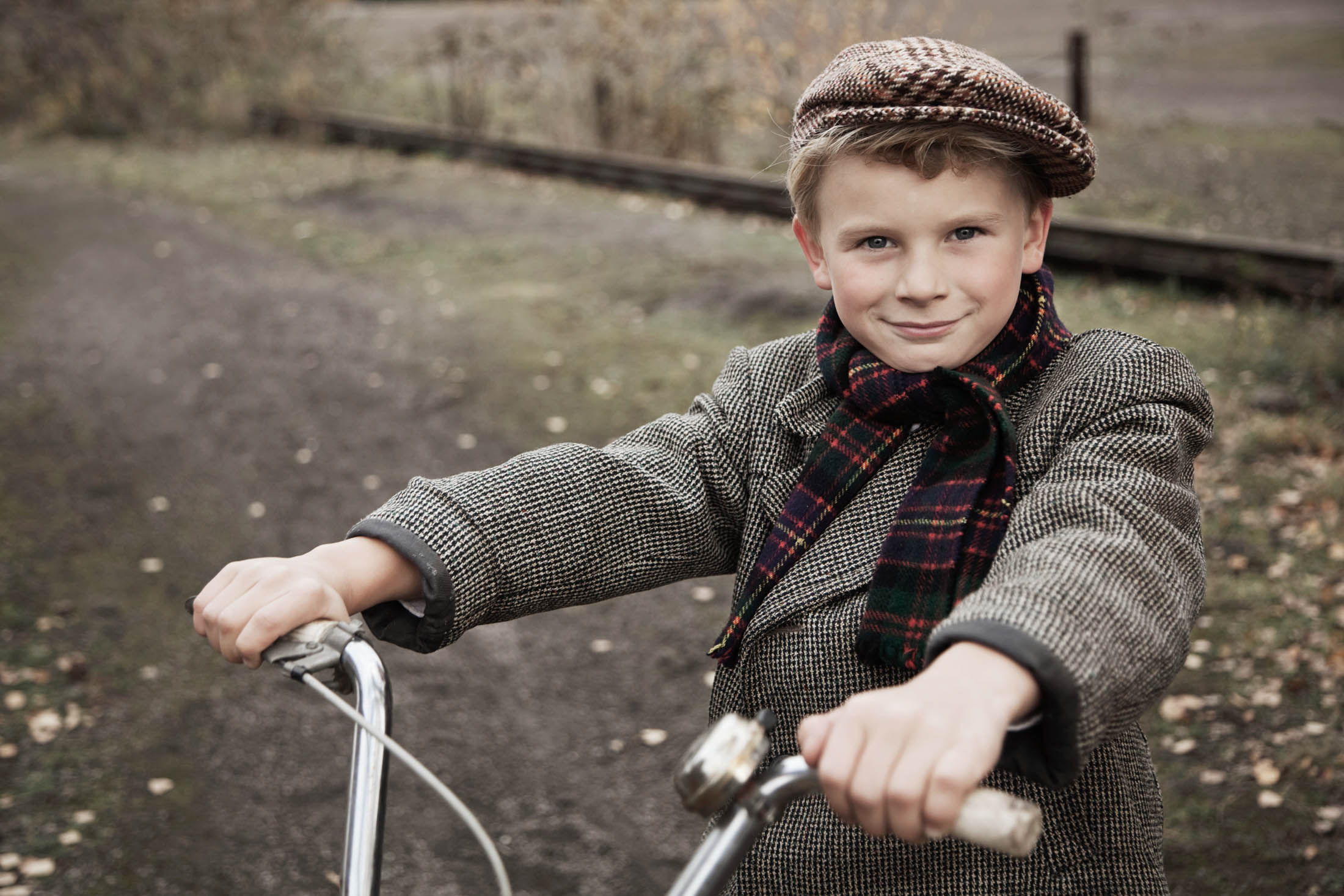 Ein kleiner Junge mit Tweedmütze und -jacke und kariertem Schal lächelt, während er den Lenker eines Fahrrads auf einem Feldweg hält, im Hintergrund sind Bäume und ein Zaun zu sehen. © Fotografie Tomas Rodriguez
