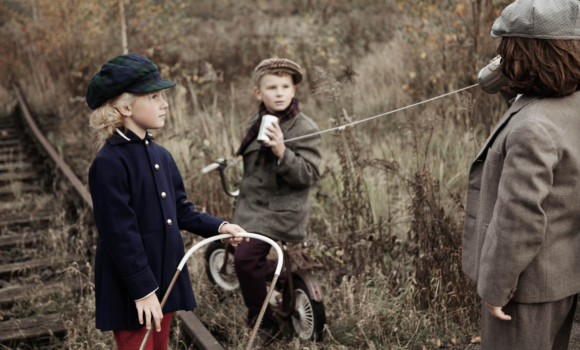 Drei Kinder in Vintage-Kleidung spielen draußen auf einer überwucherten Bahnstrecke; eines hört durch ein Blechdosentelefon zu. © Fotografie Tomas Rodriguez