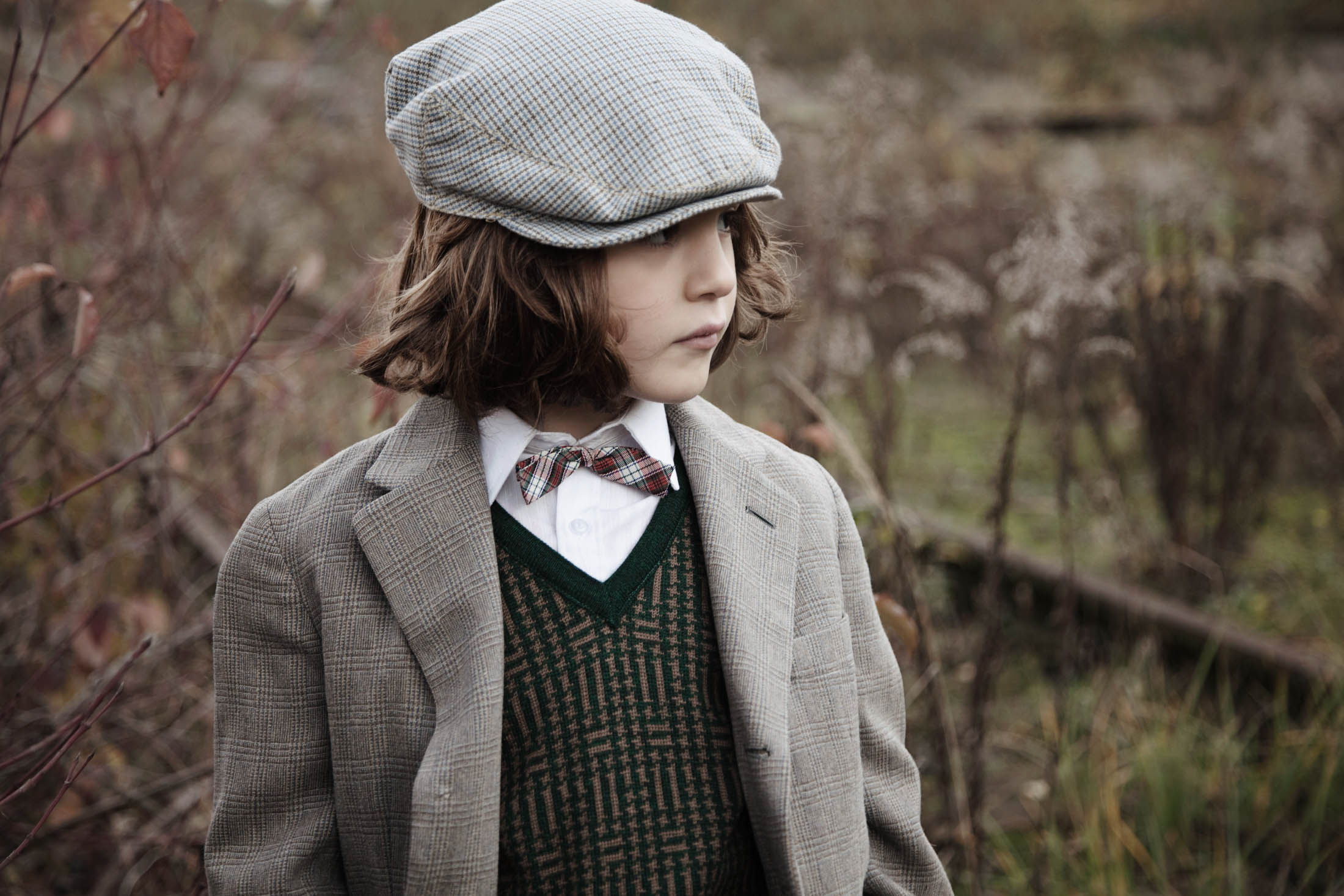 Ein kleines Kind in Vintage-Kleidung, darunter Tweedmütze, Jacke und Fliege, steht auf einem Feld mit getrockneten Pflanzen und strahlt eine nachdenkliche, altmodische Stimmung aus. © Fotografie Tomas Rodriguez