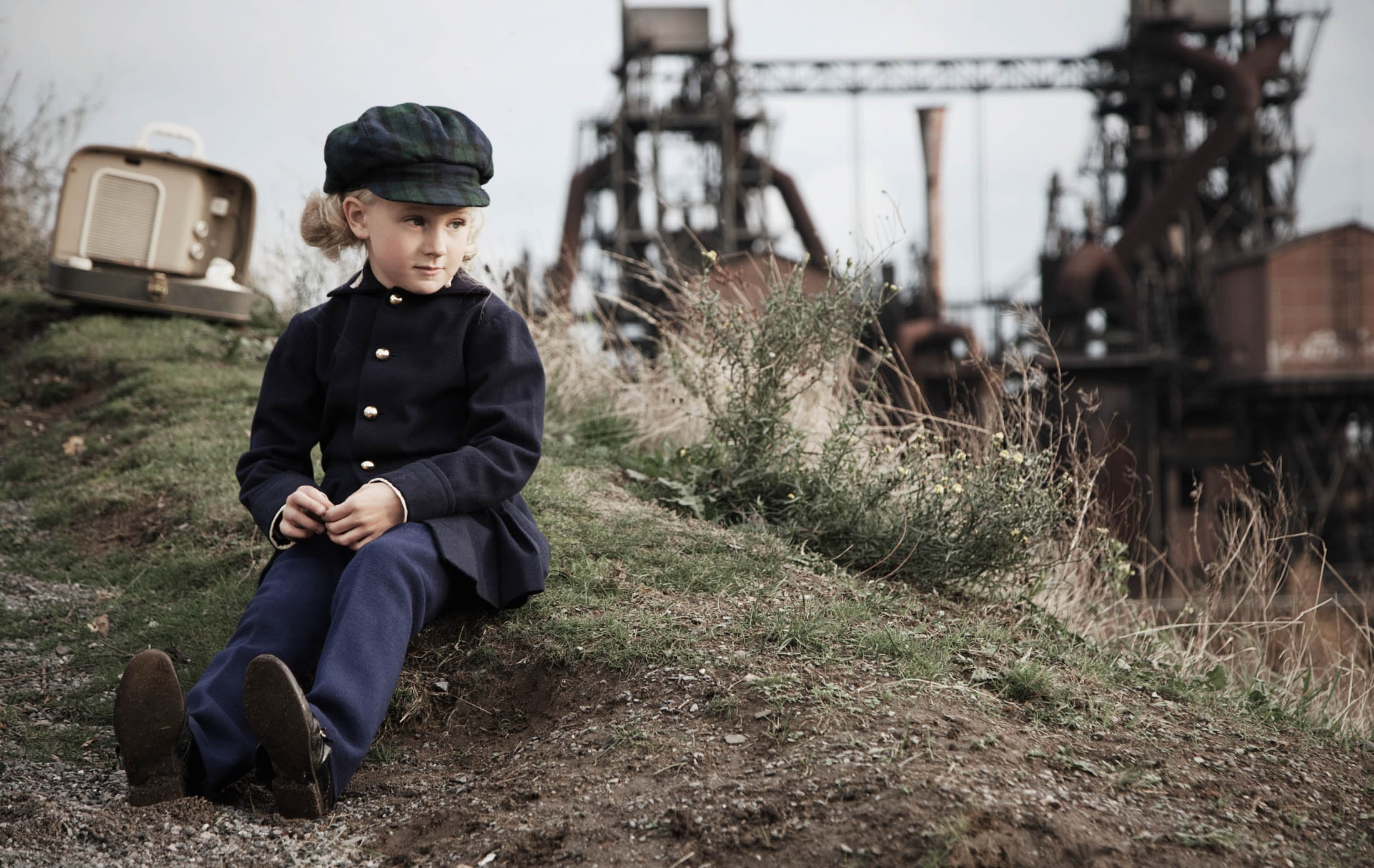 Ein kleines Kind in Vintage-Kleidung sitzt mit einem Stock in der Hand auf einem grasbewachsenen Hügel, hinter ihm steht ein alter Fernseher und in der Ferne sind Industrieanlagen zu sehen. © Fotografie Tomas Rodriguez