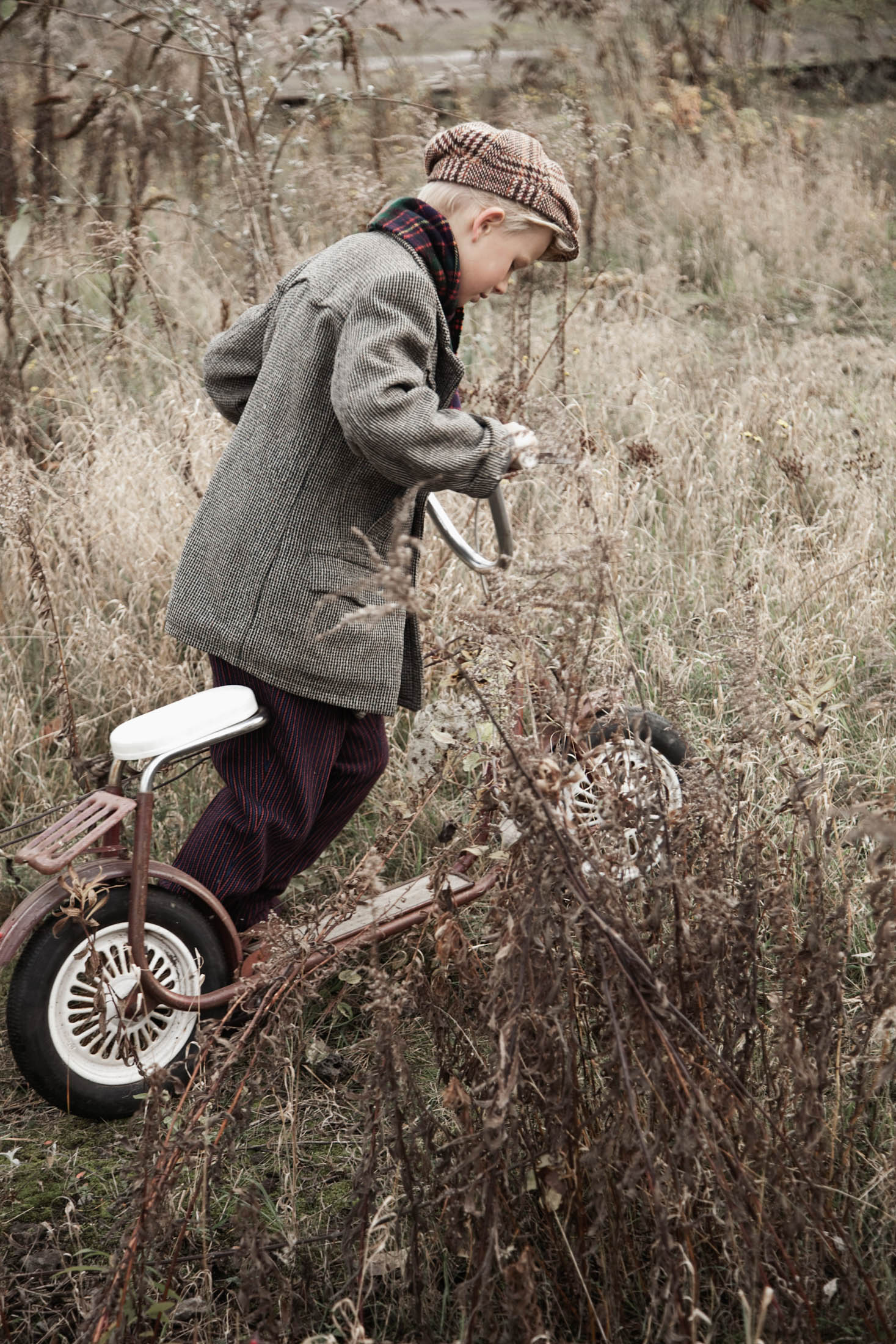 Ein kleiner Junge in Vintage-Kleidung, darunter Tweedmütze und Unterhosen, balanciert auf einem klassischen Dreirad neben überwucherten Büschen auf einem Feld und scheint etwas auf dem Boden zu untersuchen. © Fotografie Tomas Rodriguez