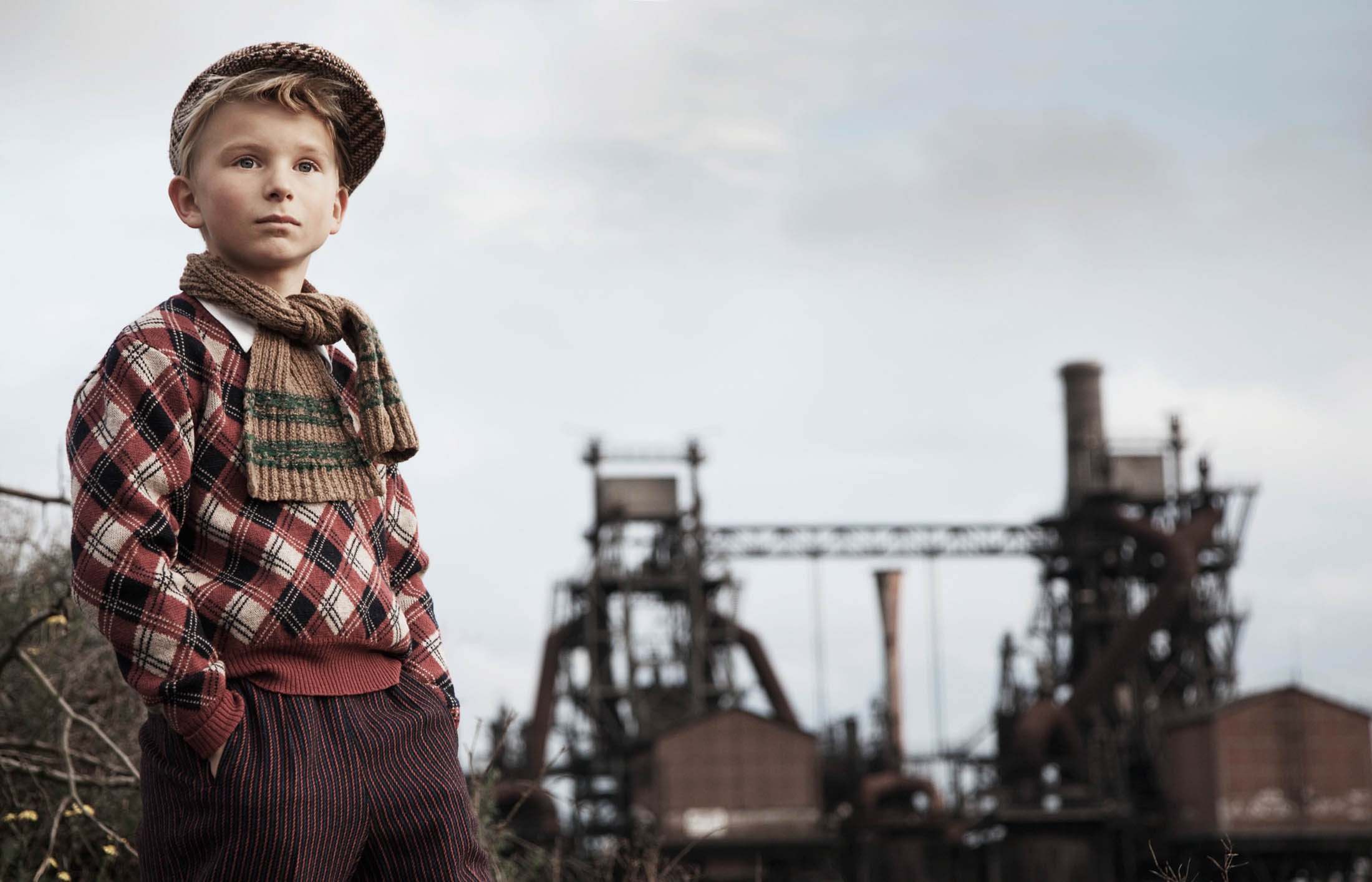 Ein kleiner Junge in Vintage-Kleidung, darunter eine karierte Jacke und eine Schiebermütze, steht selbstbewusst im Freien vor der industriellen Kulisse alter Fabriken unter einem bewölkten Himmel. © Fotografie Tomas Rodriguez