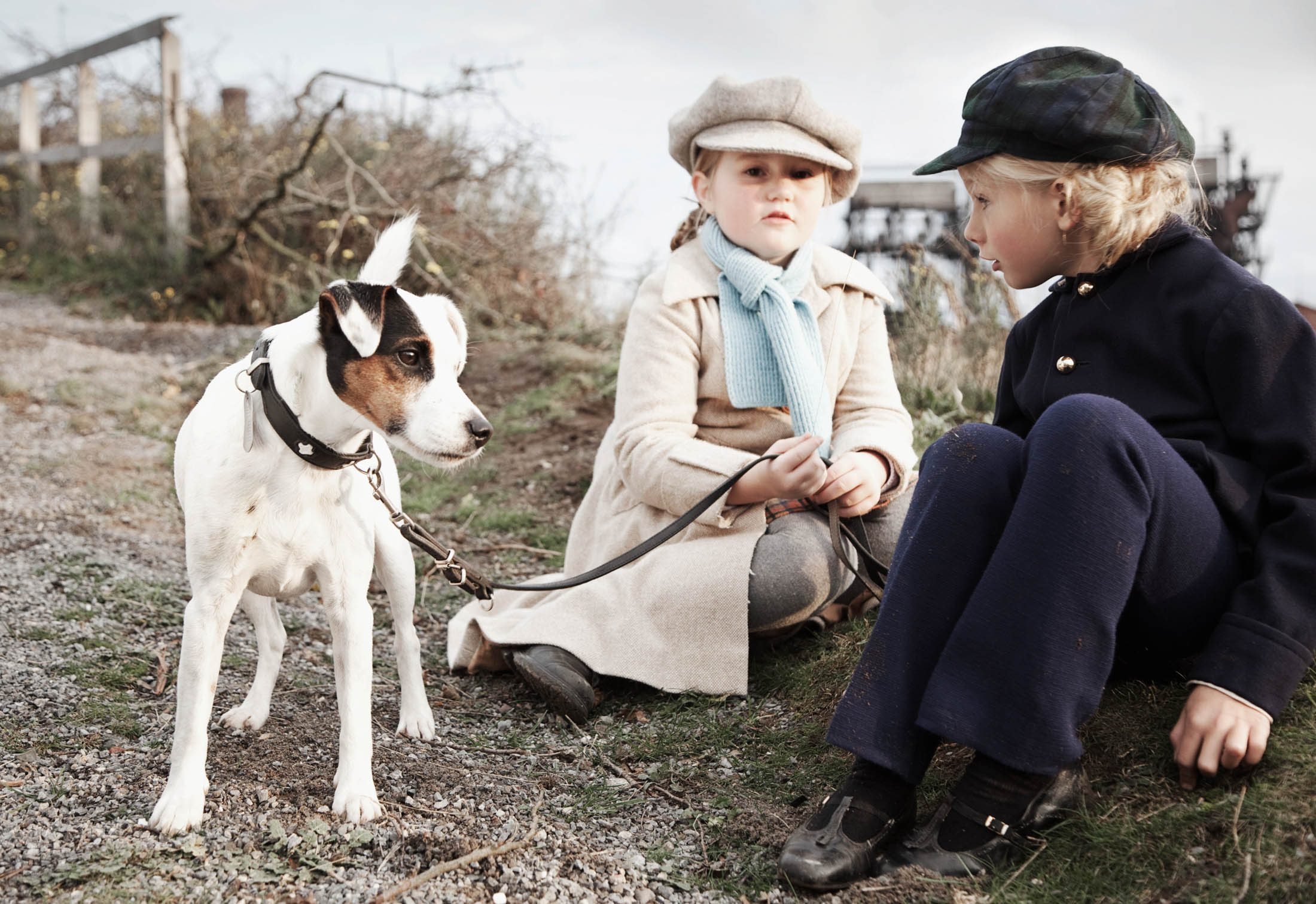 Zwei Kinder in Vintage-Kleidung sitzen im Freien, eines hält einen angeleinten Jack Russell Terrier. Die Kulisse ist rustikal und es ist ein sonniger Tag. © Fotografie Tomas Rodriguez