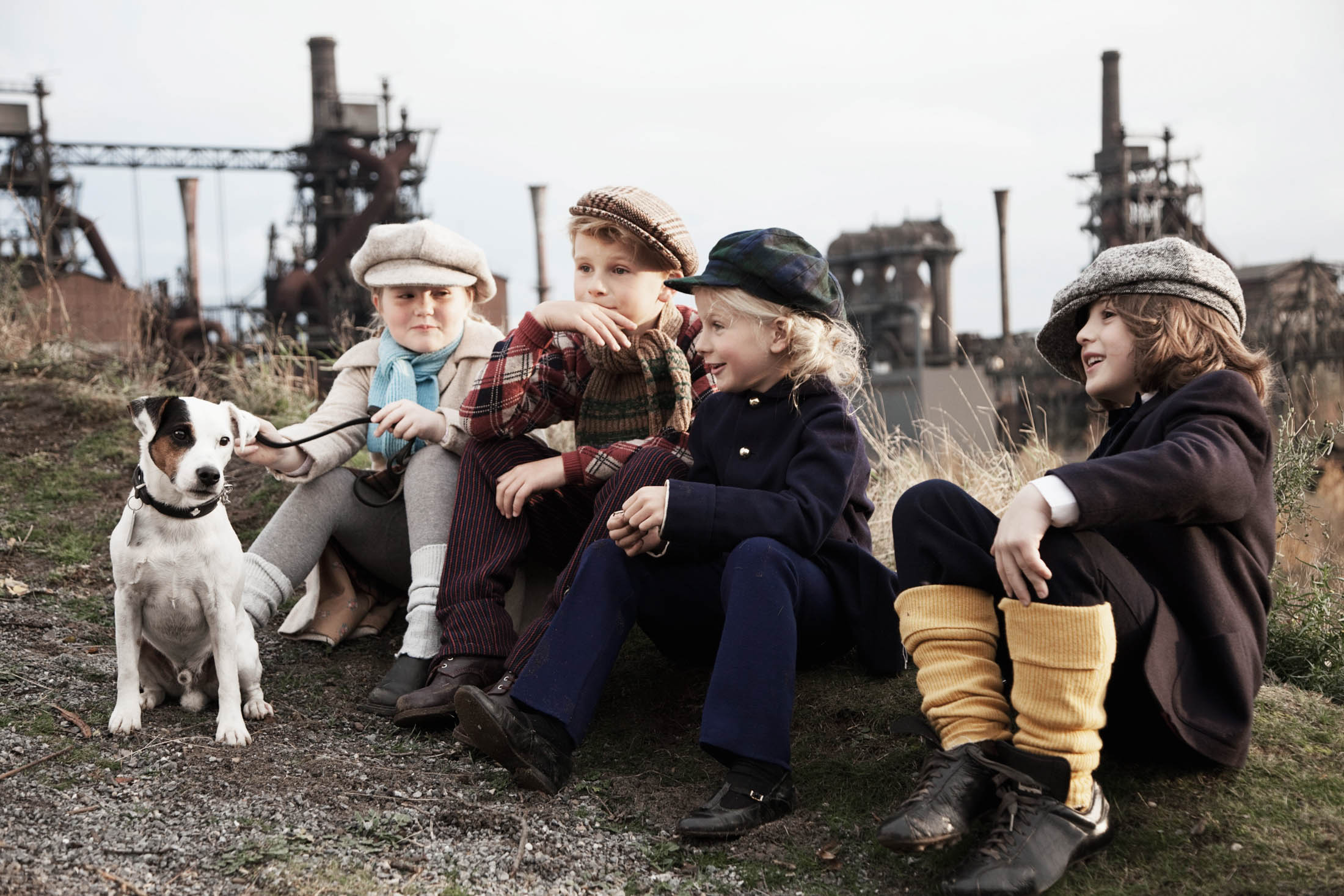 Vier Kinder in Vintage-Kleidung sitzen auf dem Boden, lachen und spielen mit einem kleinen Hund vor einer industriellen Kulisse. © Fotografie Tomas Rodriguez