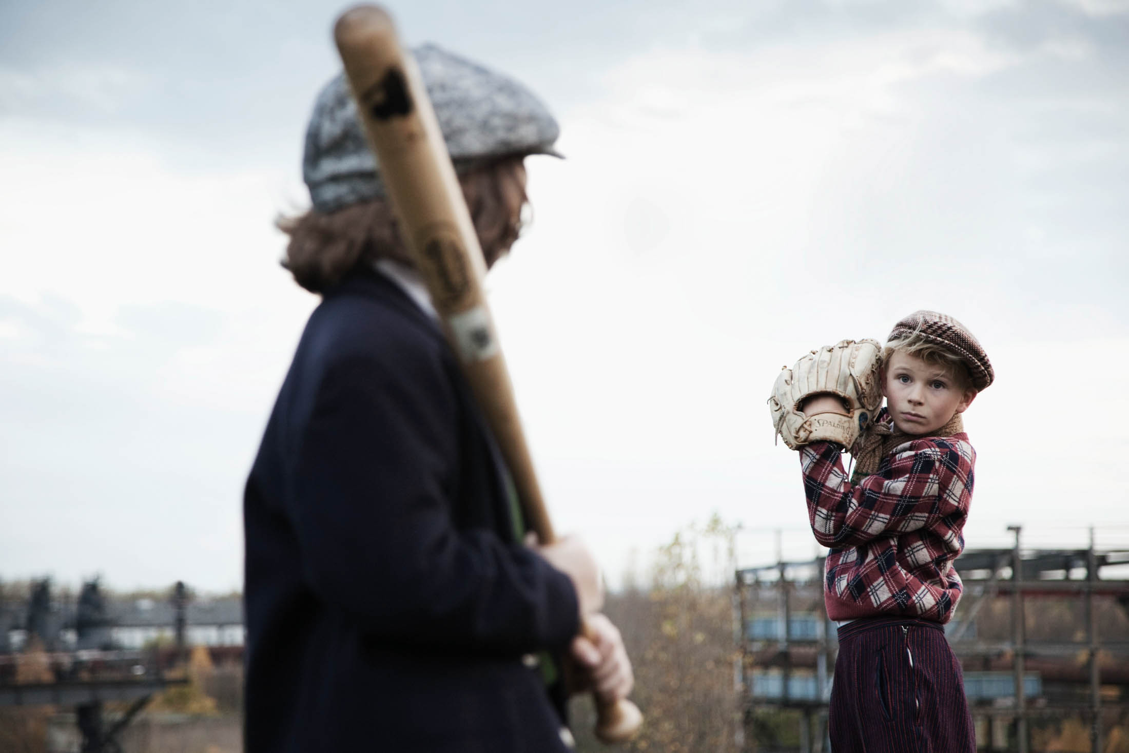 Ein kleiner Junge in kariertem Hemd und Mütze steht mit einem Baseballhandschuh bereit und beobachtet aufmerksam einen Erwachsenen, der einen Schläger hält. Beide tragen Vintage-Kleidung, im Hintergrund ist eine verschwommene Stadtlandschaft zu sehen. © Fotografie Tomas Rodriguez