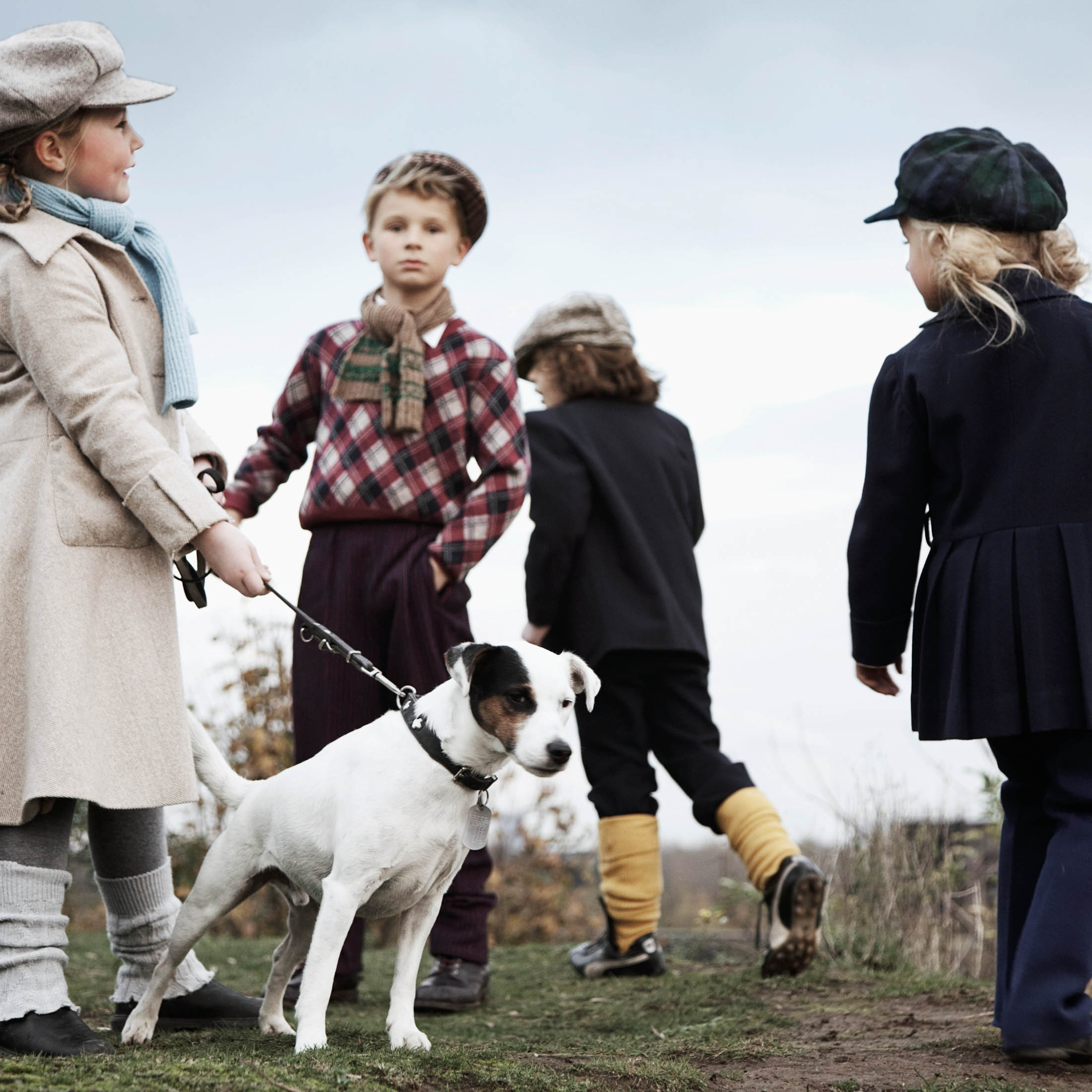 Zwei junge Mädchen und zwei Jungen in Vintage-Kleidung stehen im Freien, einer hält einen Hund an der Leine, im Hintergrund ein bewölkter Himmel. © Fotografie Tomas Rodriguez