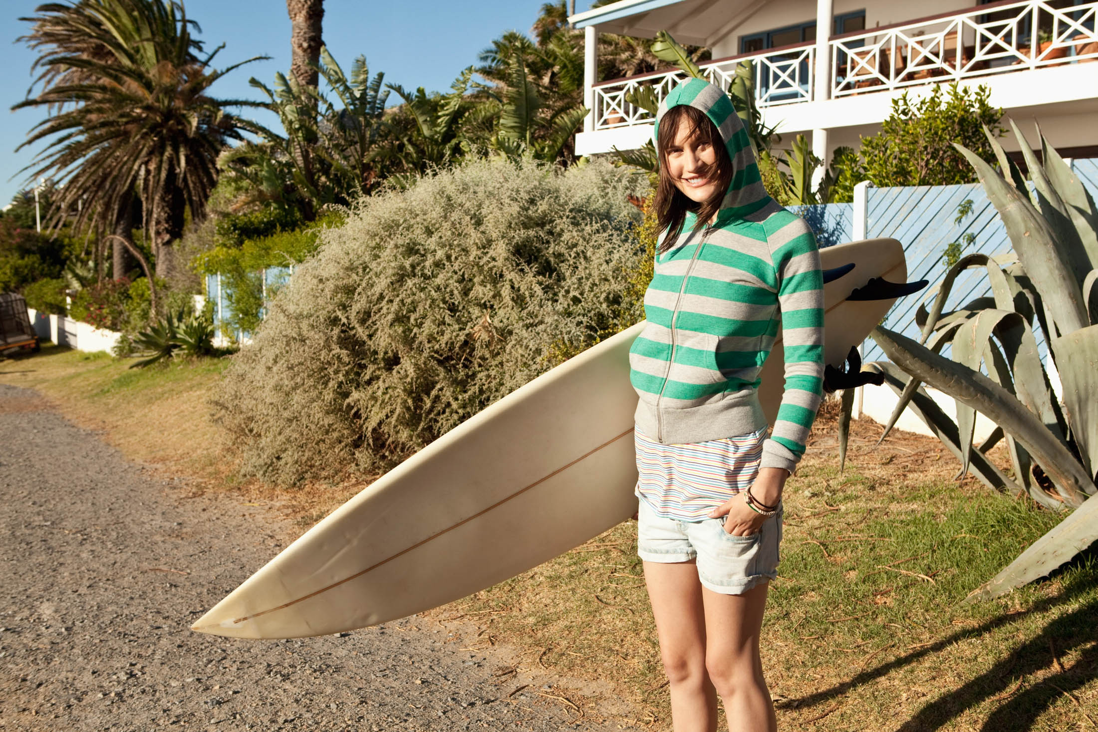 Eine Frau in einem gestreiften Kapuzenpullover und Shorts lächelt, hält ein Surfbrett unter dem Arm und steht neben tropischen Blättern und einem Haus mit Balkon. © Fotografie Tomas Rodriguez