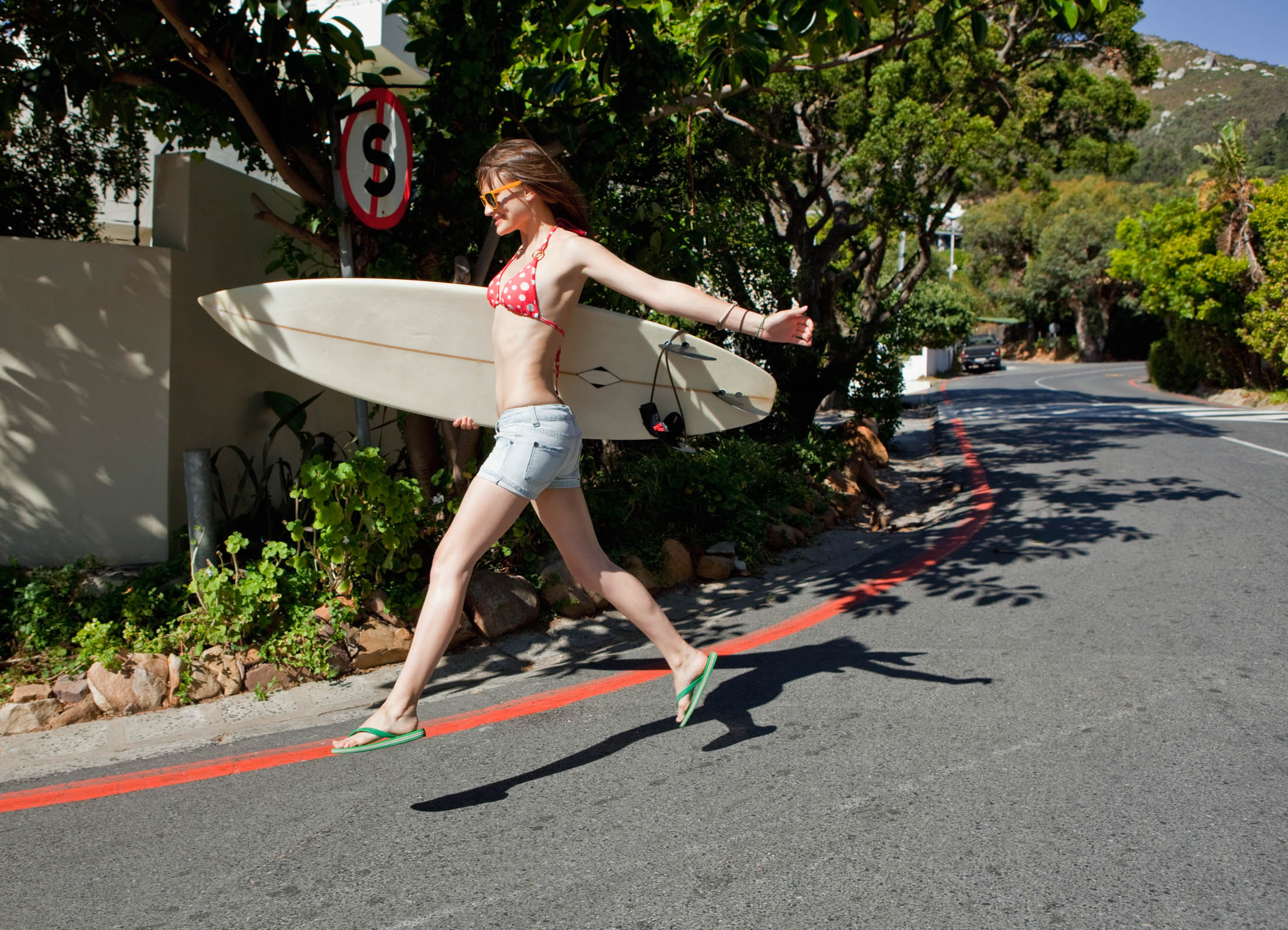 Eine junge Frau in einem roten Bikinioberteil und Jeansshorts trägt ein Surfbrett, während sie in der Nähe eines Stoppschildes eine Straße überquert, im Hintergrund ist üppiges Grün zu sehen. © Fotografie Tomas Rodriguez